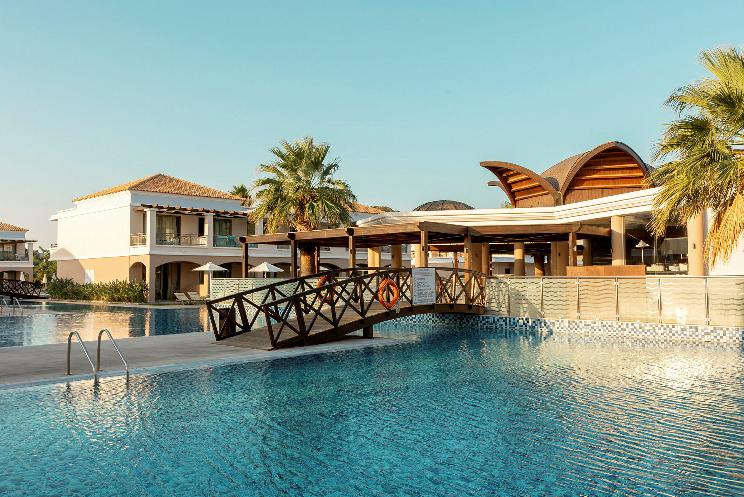 Utsikt över en hotellpool med en bro och byggnader i bakgrunden. (AI-genererad text)