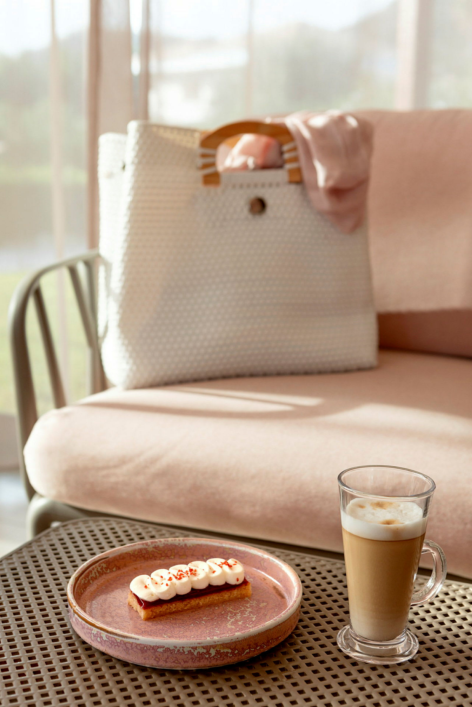 En beige väska med trähandtag på en rosa sittmöbel med en glas med latte och en dessert på en tallrik i förgrunden. (AI-genererad text)