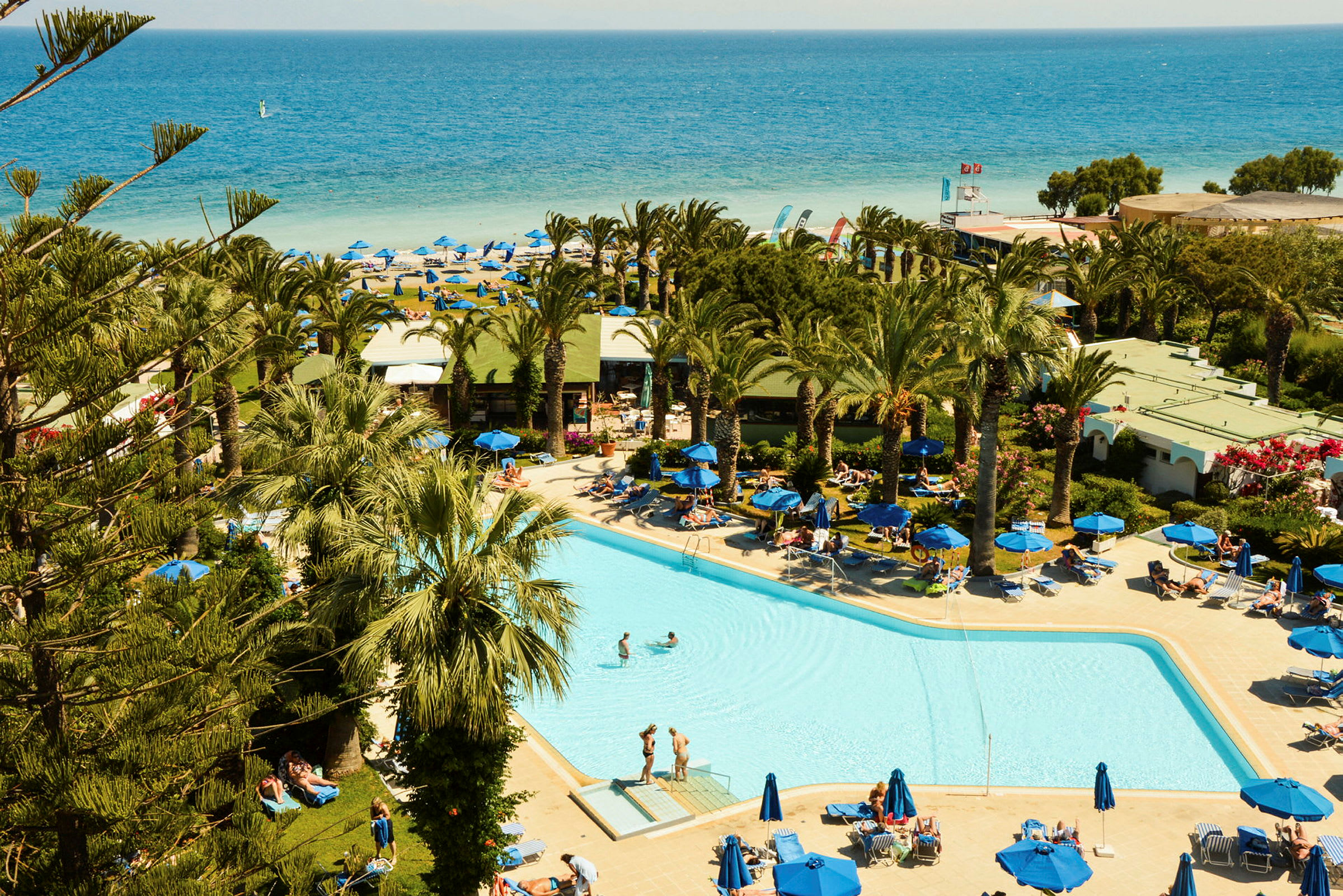 En pool omgiven av solstolar och palmer, med en sandstrand och havet i bakgrunden. (AI-genererad text)