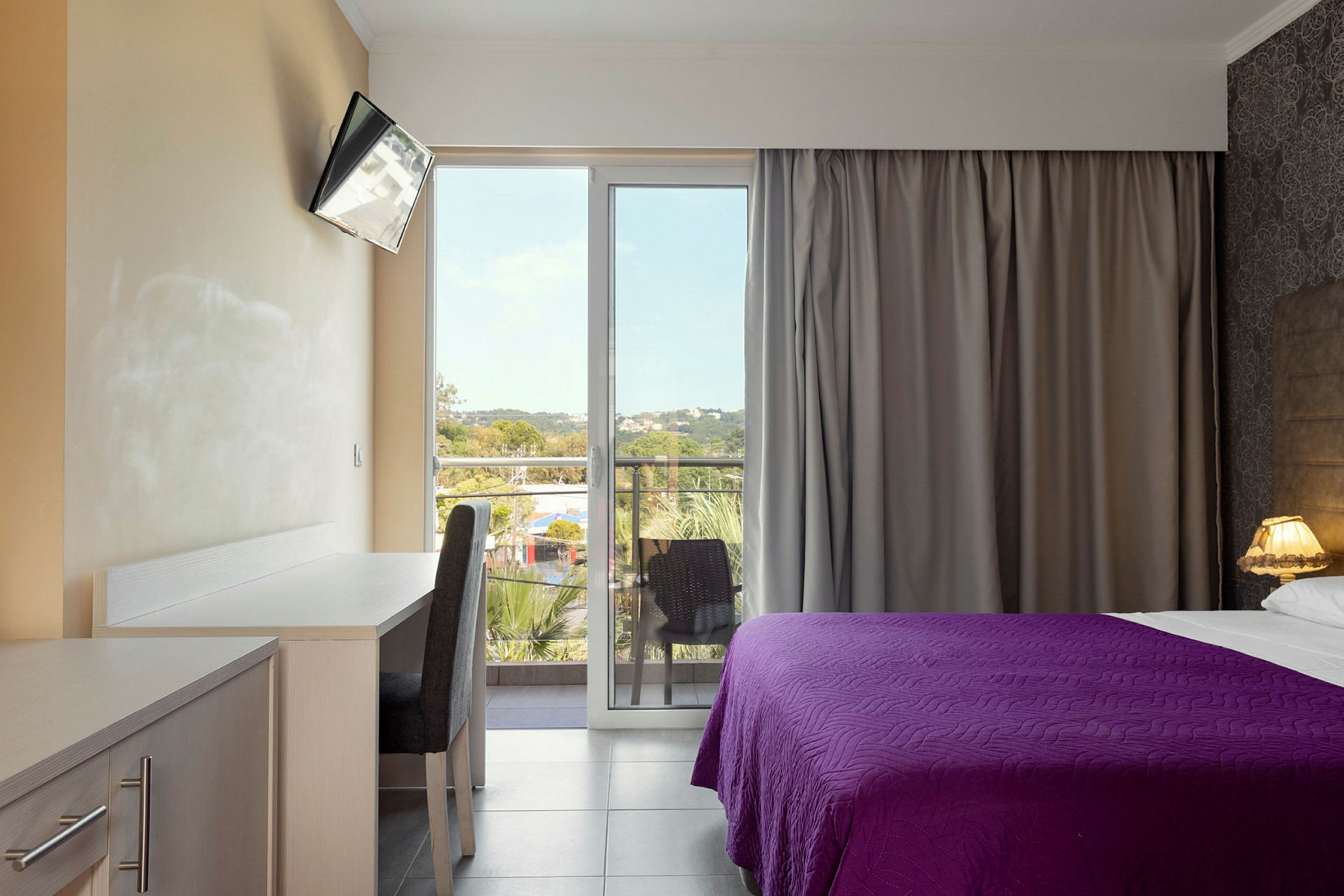 Ett hotellrum med ett skrivbord, en stol, en TV och en säng med lila överkast. Rummet har en balkong med utsikt. (AI-genererad text)