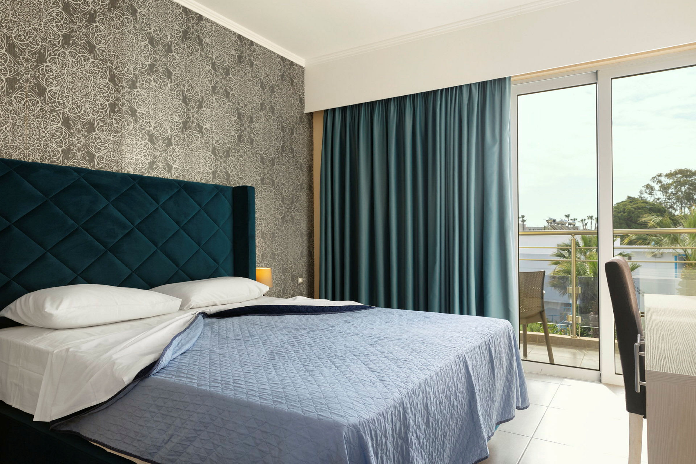 Ett hotellrum med en säng, blått överkast och blå gardiner, samt ett fönster som leder till en balkong. (AI-genererad text)