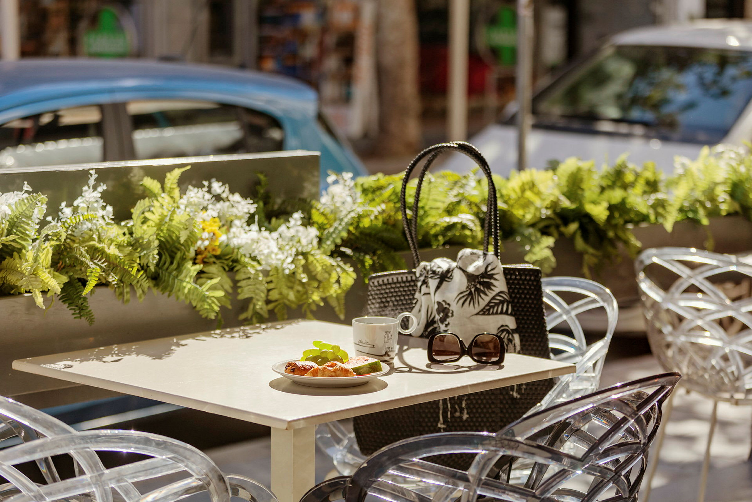 Bord med croissant, kopp och solglasögon på uteservering med blomsterdekorationer. (AI-genererad text)