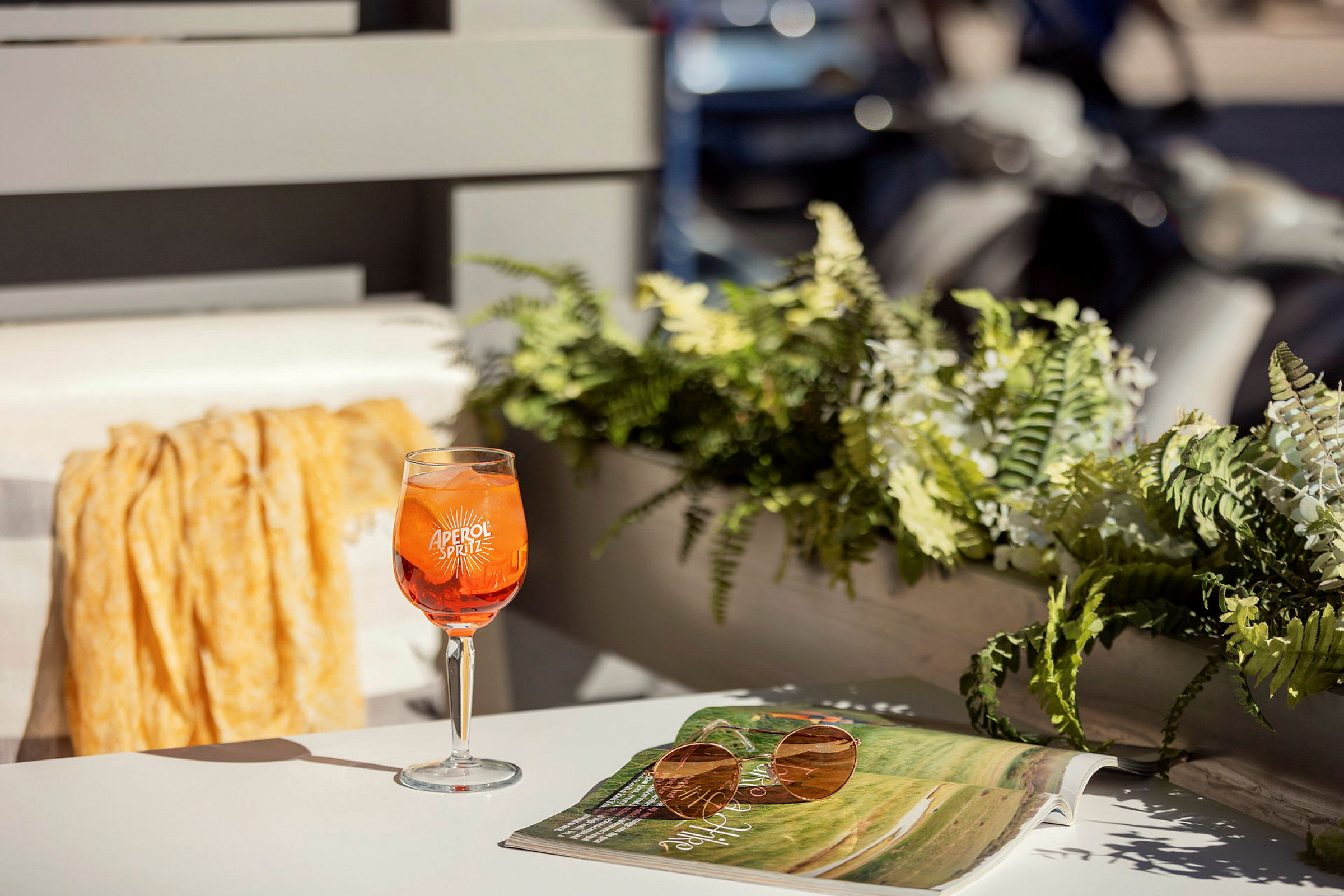 Ett glas Aperol Spritz står på ett bord bredvid en solglasögon och en öppen tidning. I bakgrunden syns växter och delar av en balkong. (AI-genererad text)