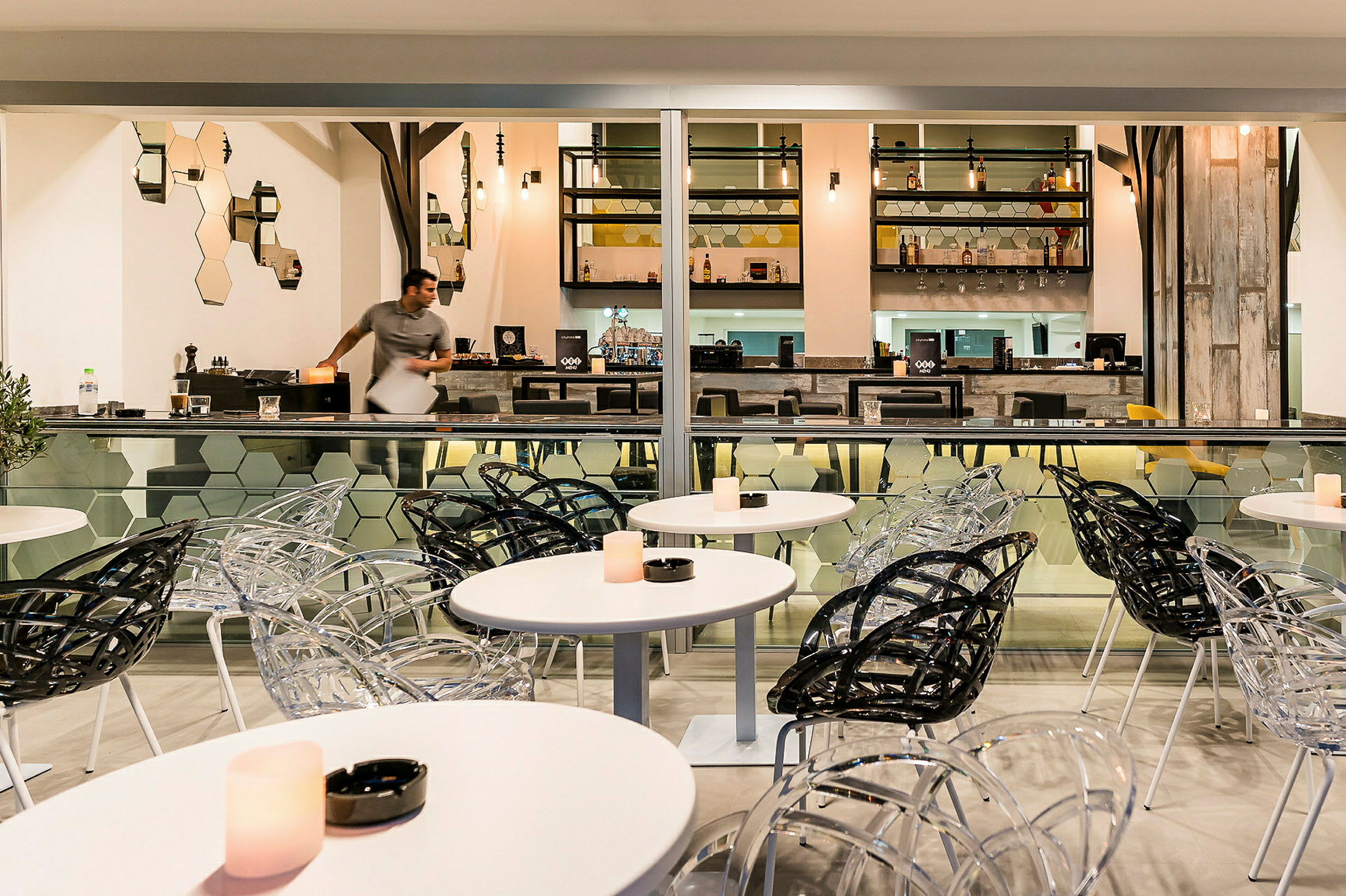 Ett modernt café med genomskinliga och svarta stolar samt vita bord. Bakom finns en välbelyst bar. (AI-genererad text)