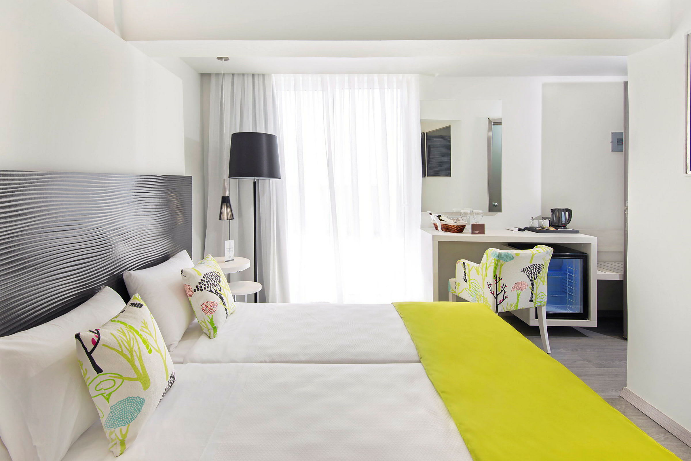 Ett ljust hotellrum med två enkelsängar, dekorativa kuddar och en limegrön sängfilt, samt en sittplats och ett skrivbord med spegel. (AI-genererad text)