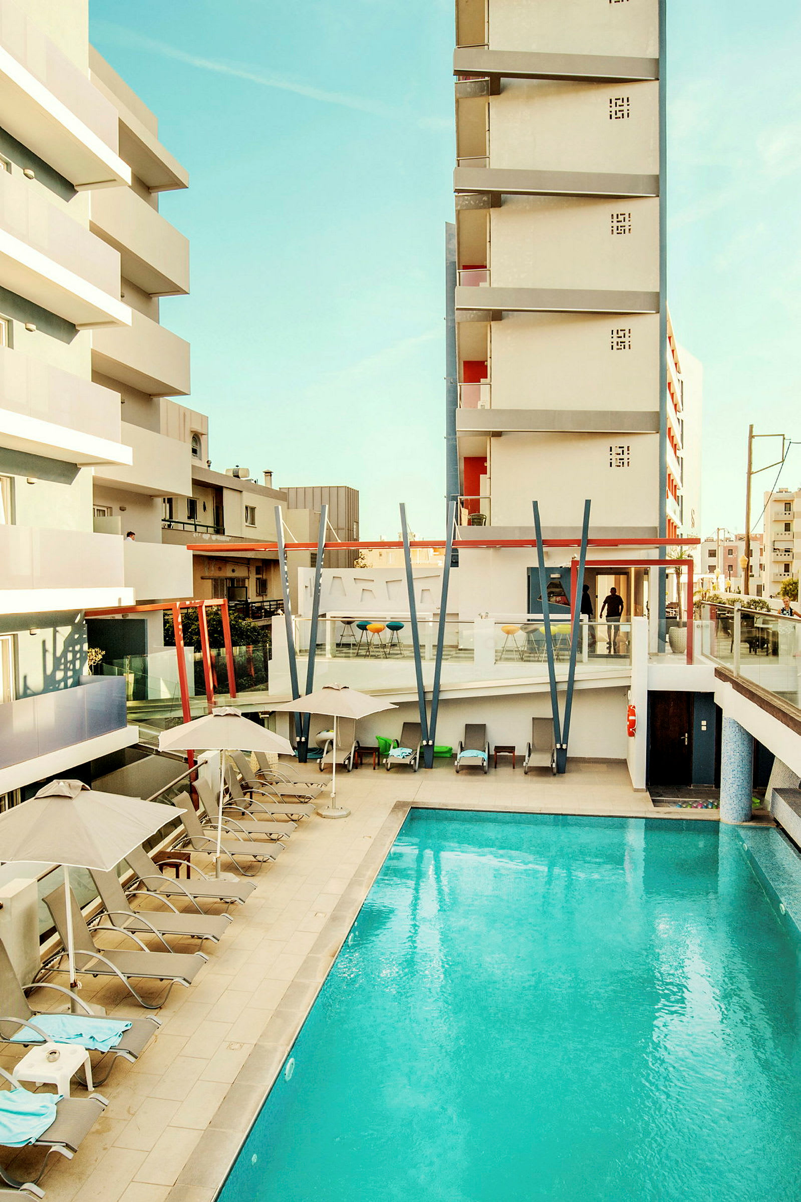 Bild av ett hotellområde med pool och solstolar, omgiven av byggnader. (AI-genererad text)