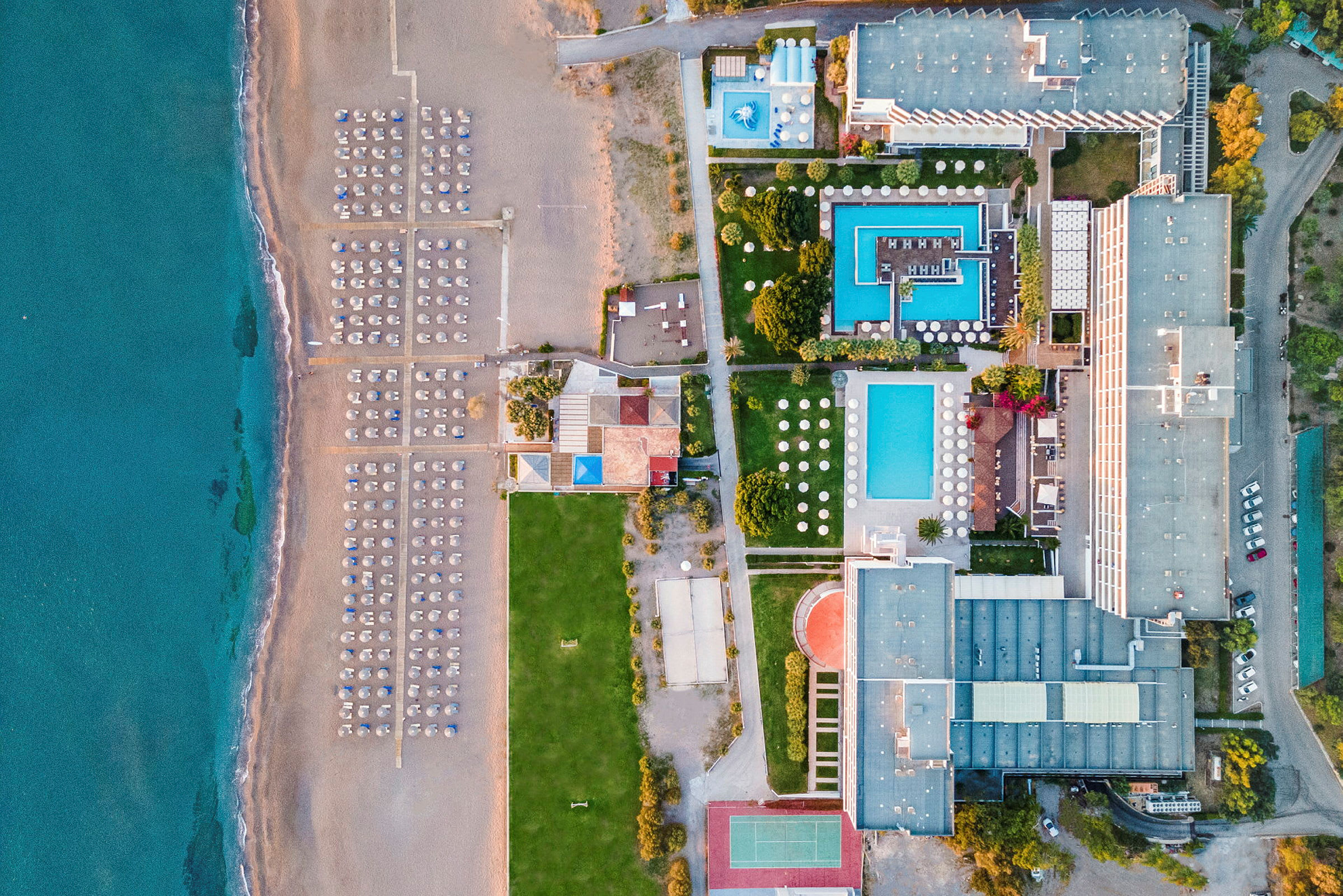 Flygfoto över en strand med solstolar och parasoller vid havet samt ett hotellområde med pooler och byggnader. (AI-genererad text)