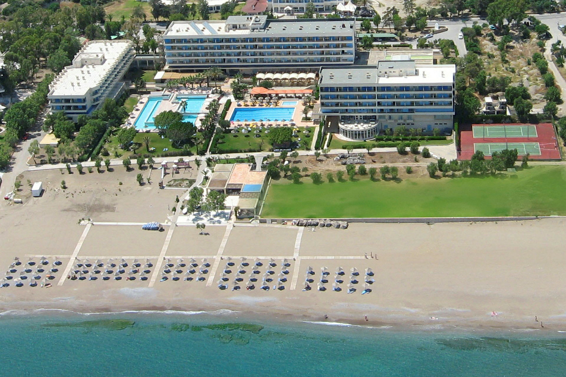 Luftvy av ett hotellkomplex med pooler, grönområden och tennisbanor nära en sandstrand. (AI-genererad text)
