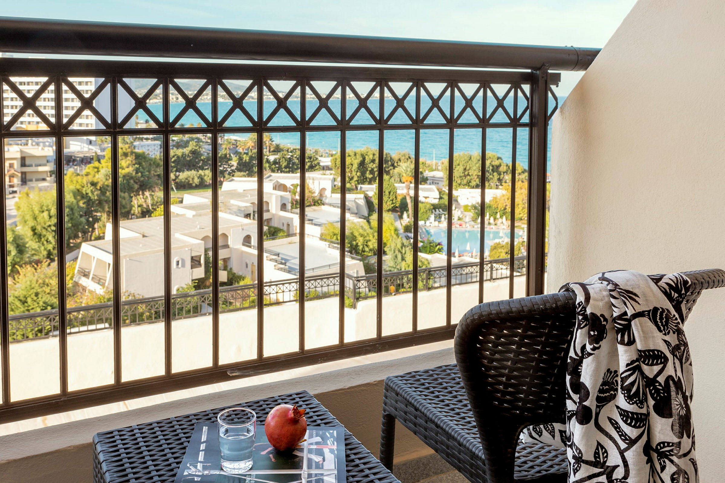 En balkong med utsikt över hotellområdet och havet. På balkongens bord står ett glas vatten och ett granatäpple. På en stol ligger en mönstrad sjal. (AI-genererad text)