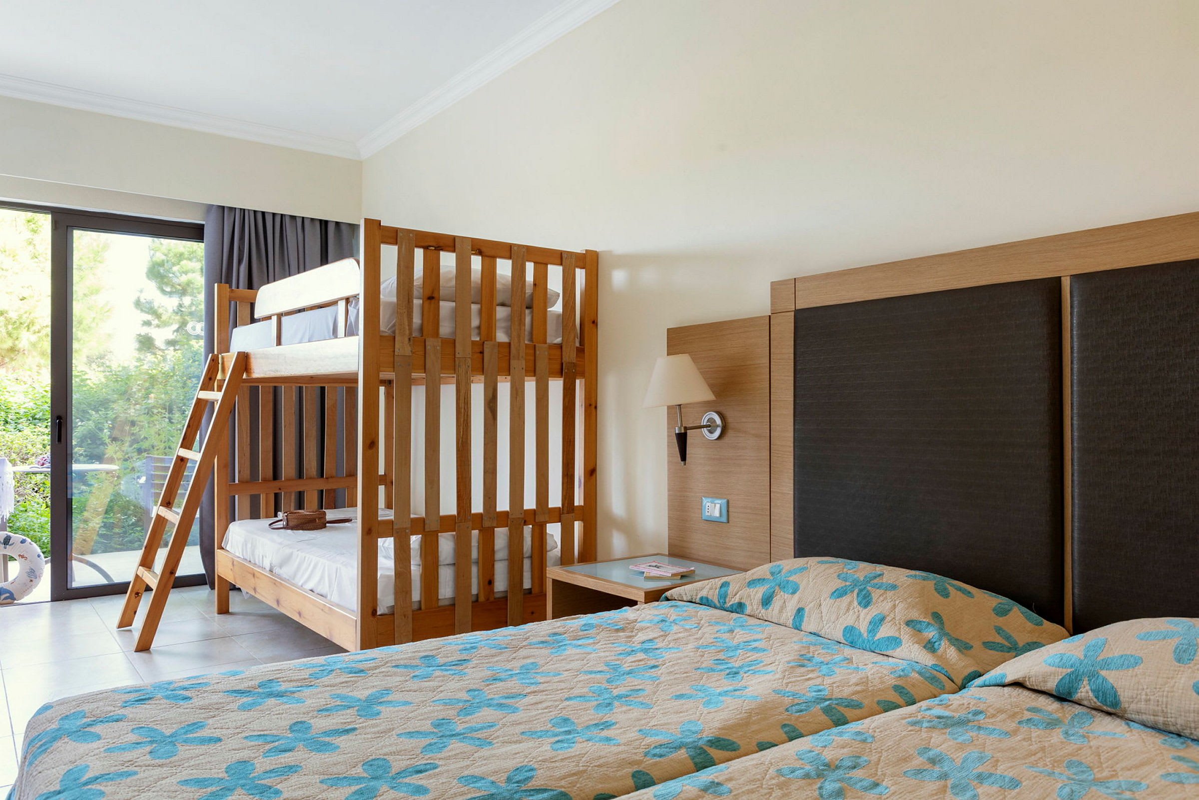 Ett hotellrum med två enkelsängar med blommiga överkast och en våningssäng i trä bredvid ett fönster. (AI-genererad text)