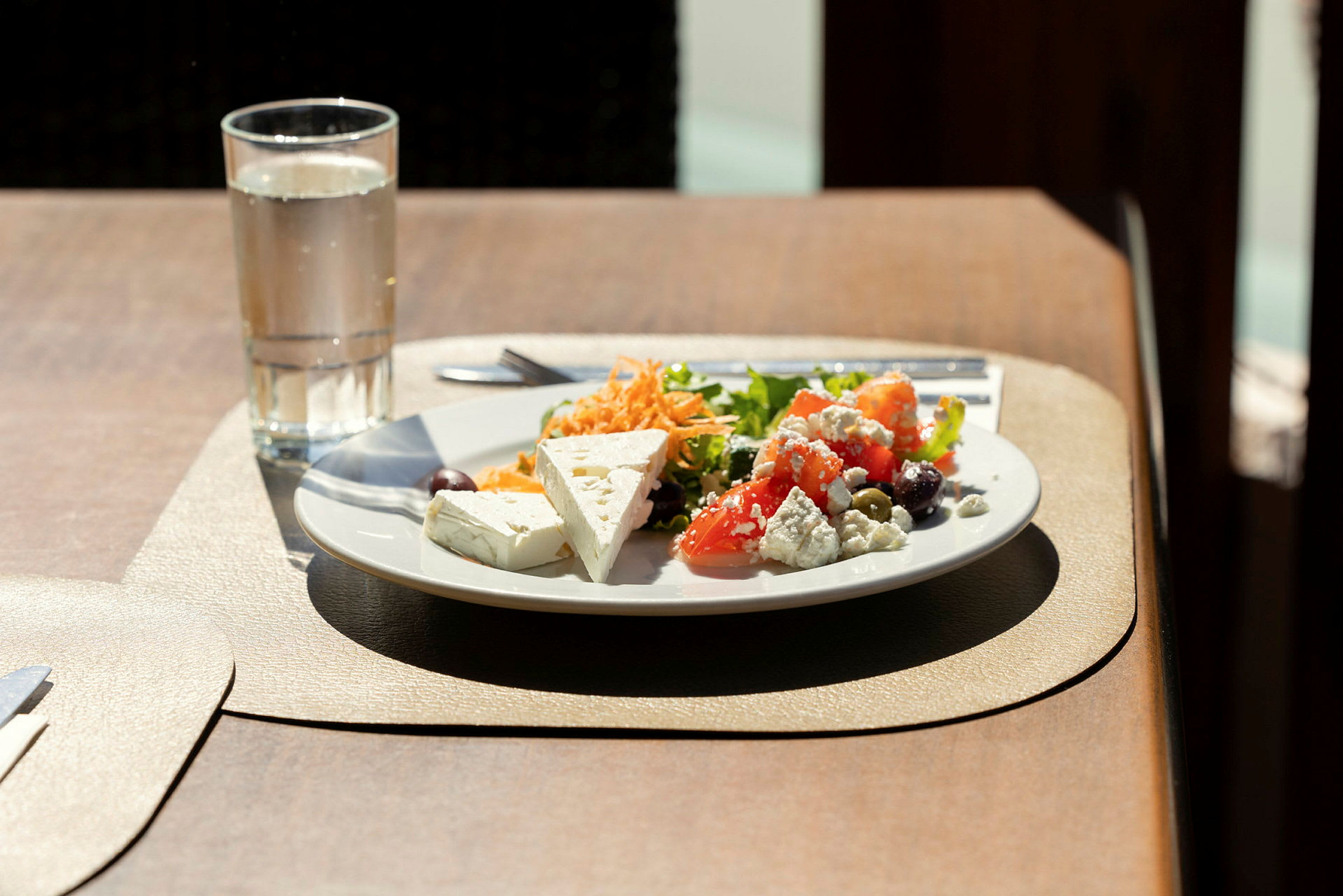 En tallrik med sallad, grönsaker, och fetaost samt ett glas vatten på ett bord. (AI-genererad text)