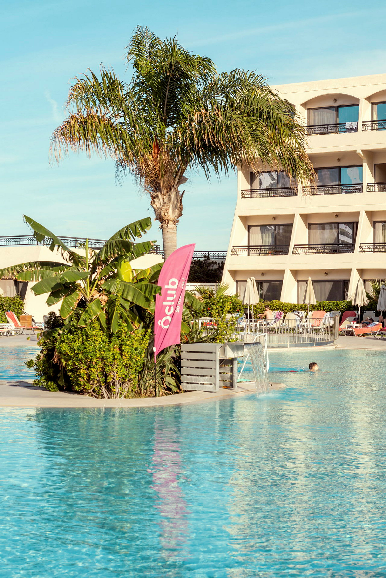 Utomhuspool framför hotellbyggnad med palmer och växtlighet. (AI-genererad text)