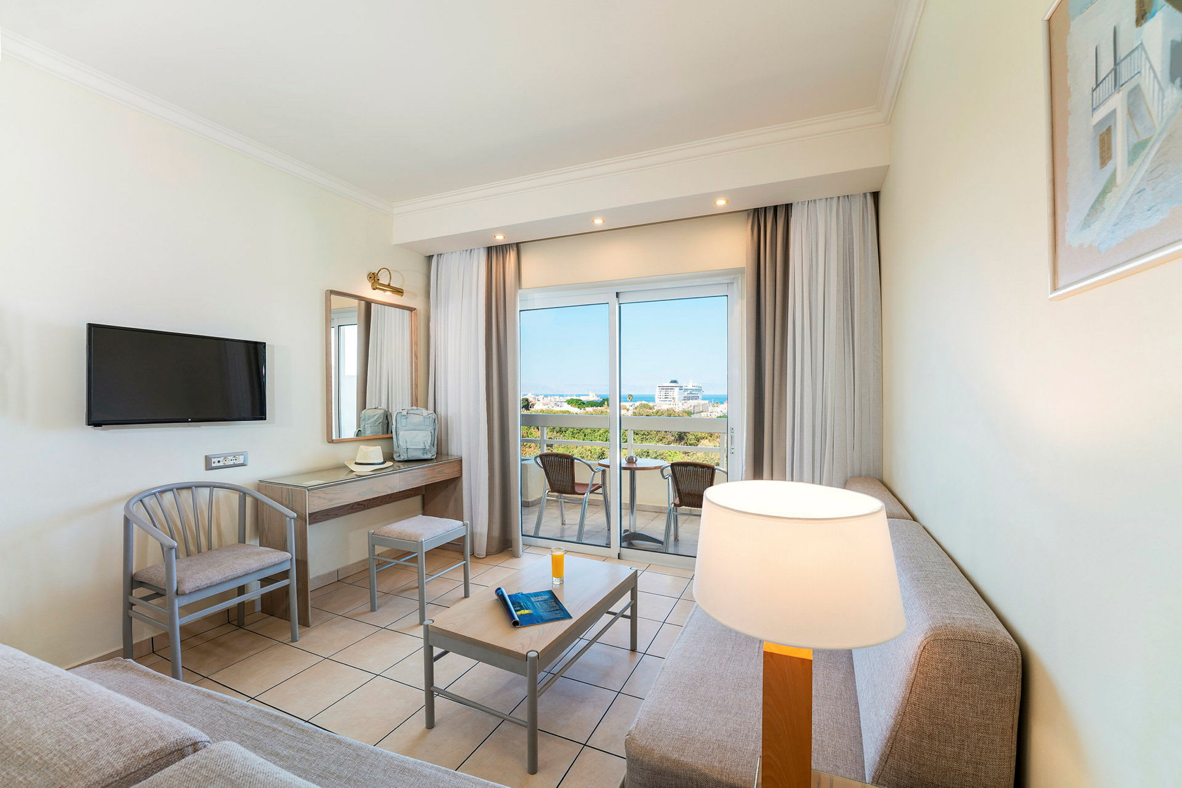 Ett hotellrum med sittgrupp och bord, en TV på väggen, ett skrivbord och en balkong med utsikt. (AI-genererad text)