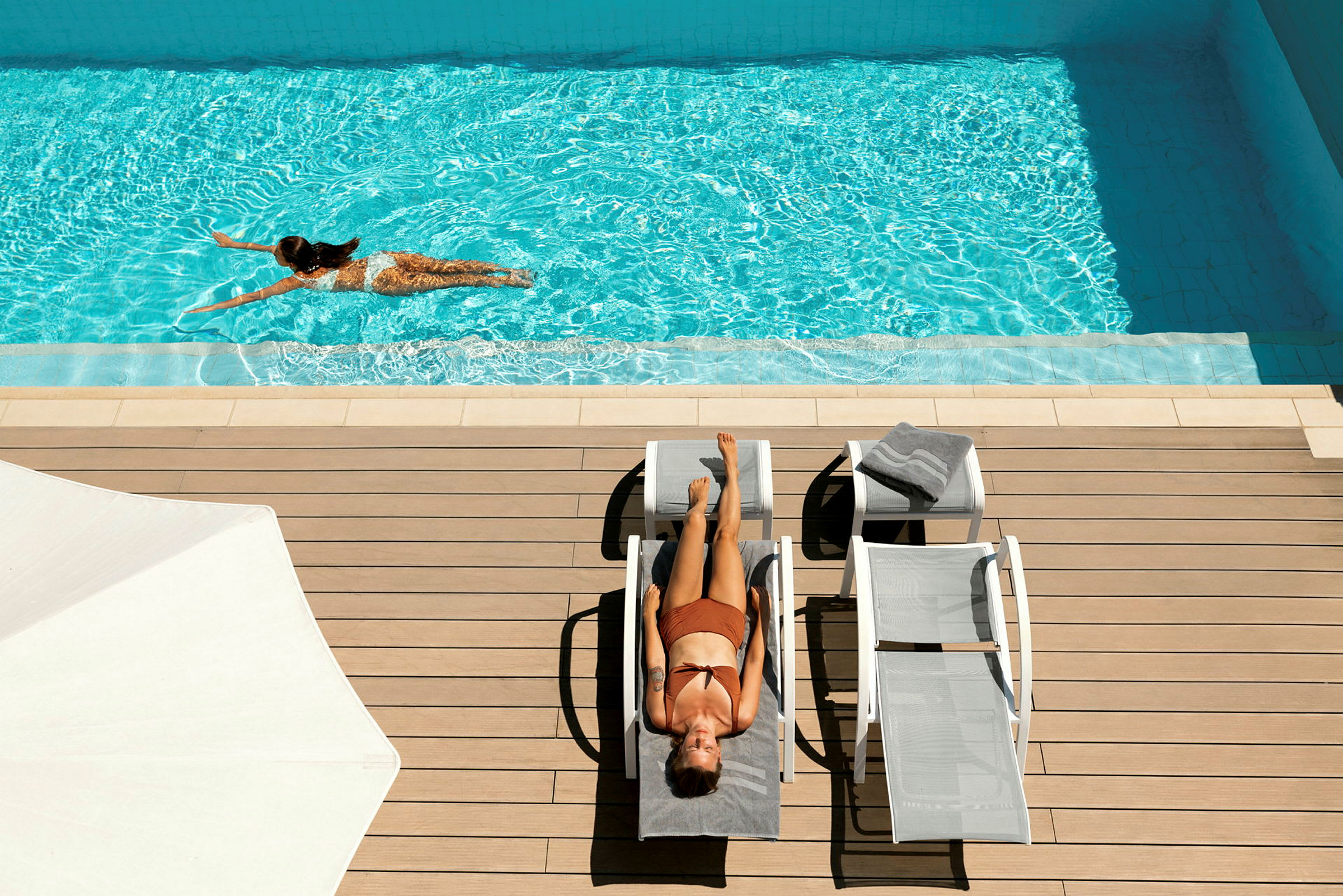 En kvinna ligger och solar på en solstol vid en pool. En annan kvinna simmar i poolen. (AI-genererad text)