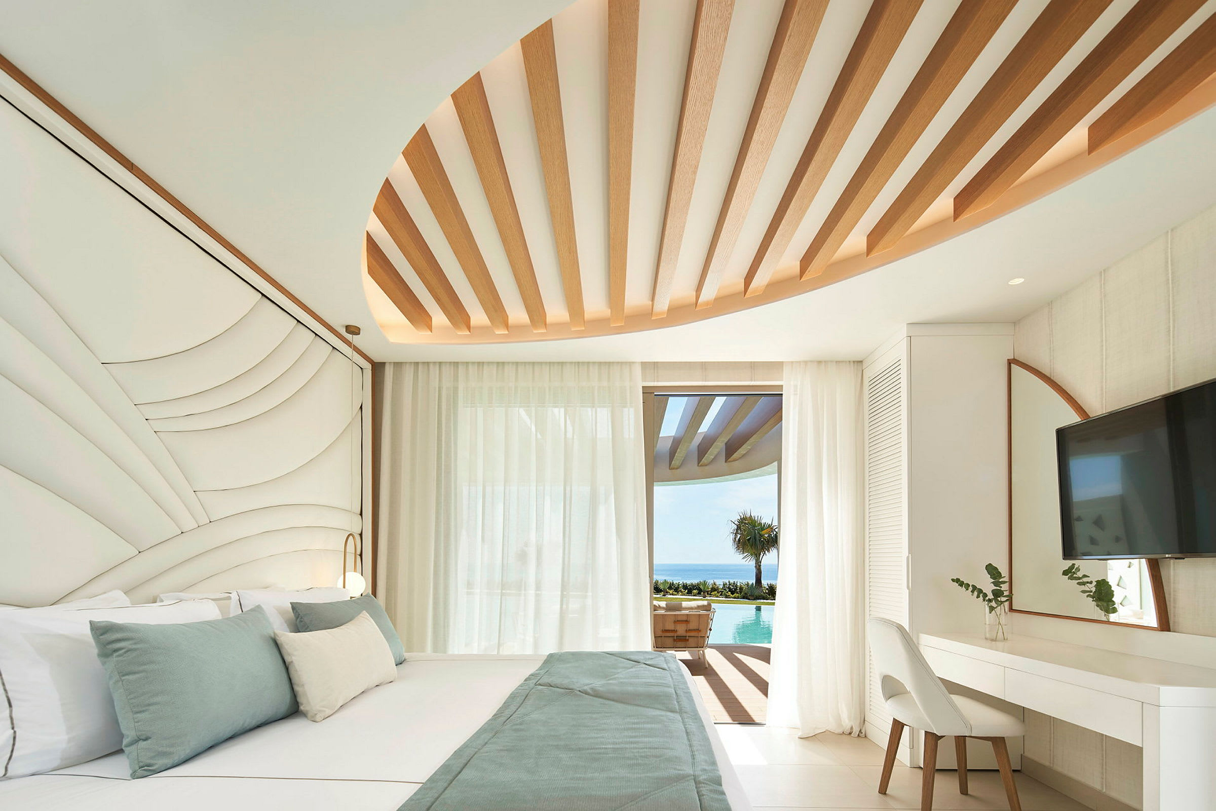 Ett modernt hotellrum med en stor säng, designade takdetaljer och utsikt över en pool och havet genom en glasdörr. (AI-genererad text)
