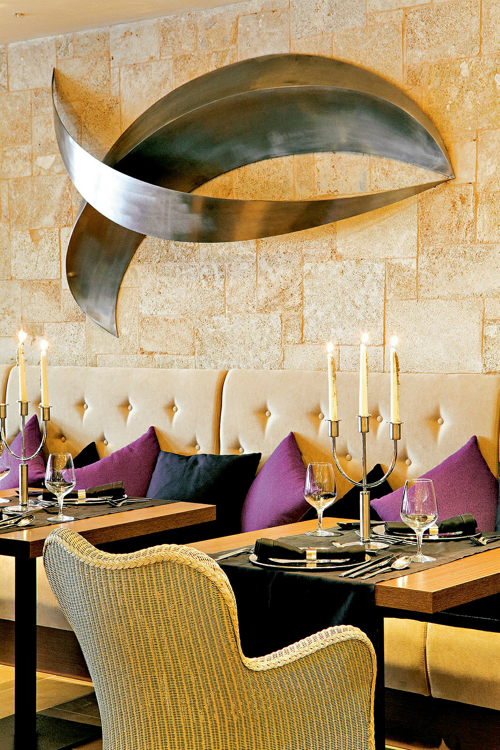 Ett elegant restaurangbord med tallrikar, glas och ljusstakar, omgiven av stoppade soffor och färgade kuddar, och en modern konstinstallation på väggen. (AI-genererad text)