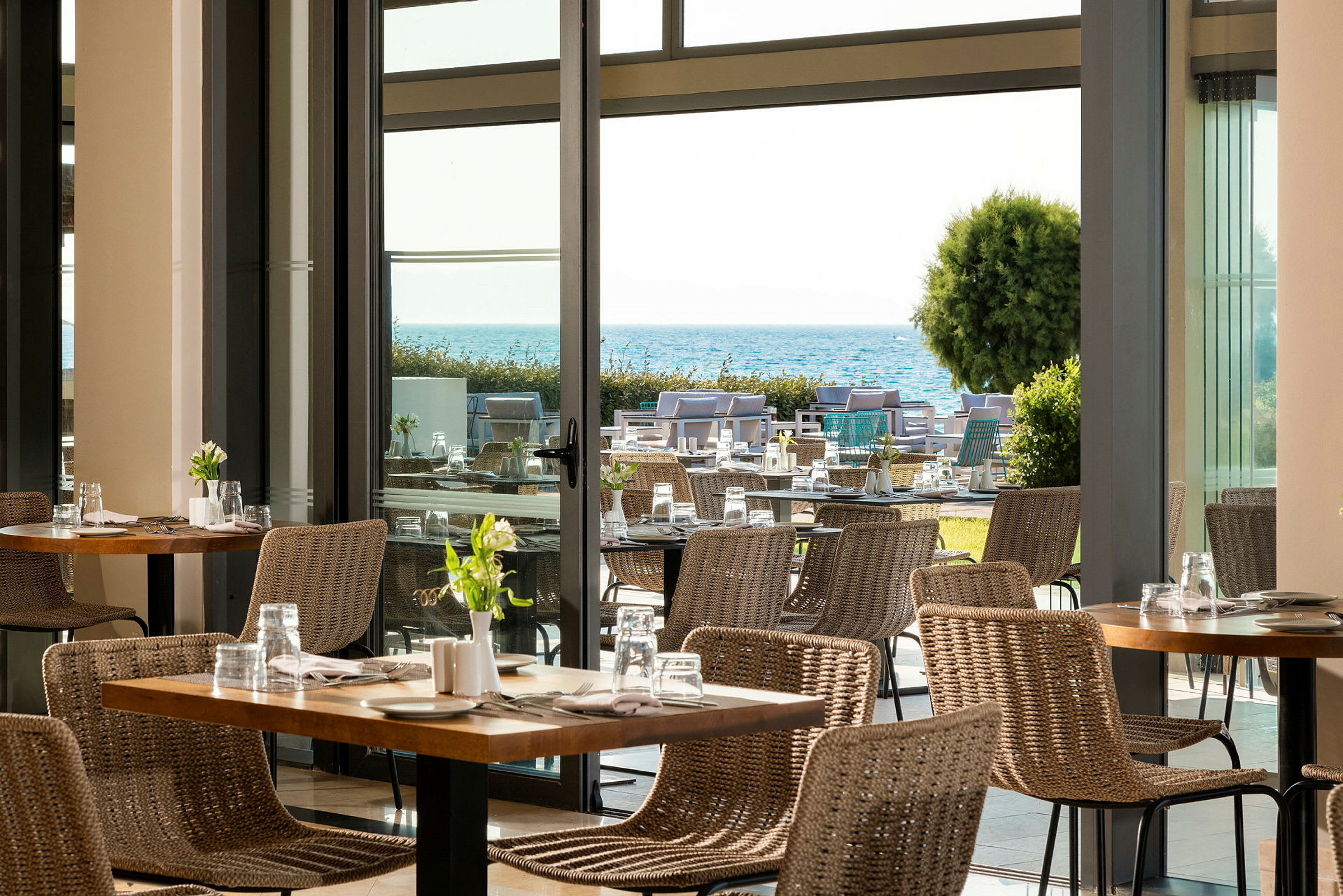Utsikt över en modern restaurang med rottingstolar och bord, med en trädgård och havet i bakgrunden. (AI-genererad text)