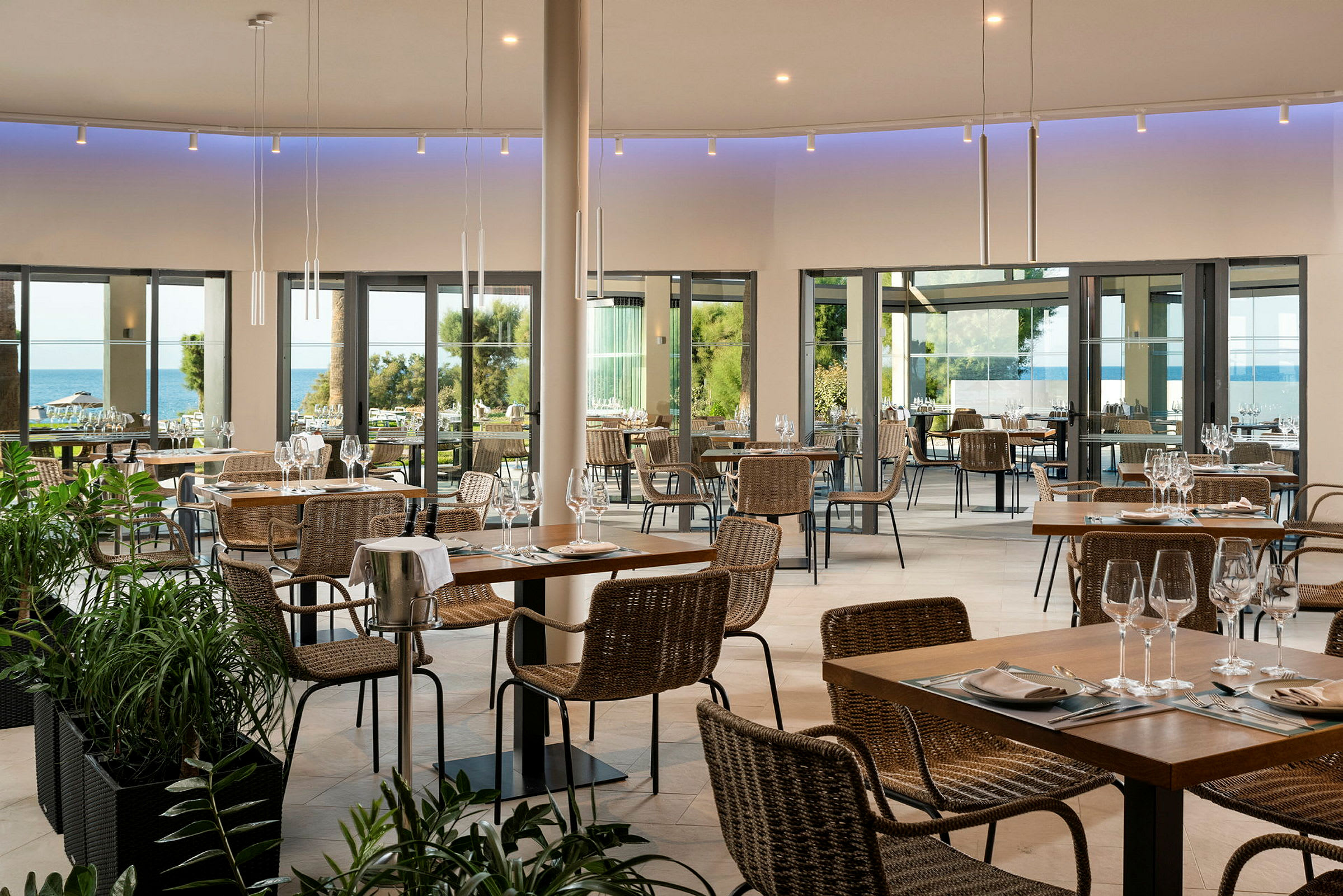 En restaurang med bord och stolar, stora glasfönster som vetter mot en utsikt av havet och grönska. (AI-genererad text)