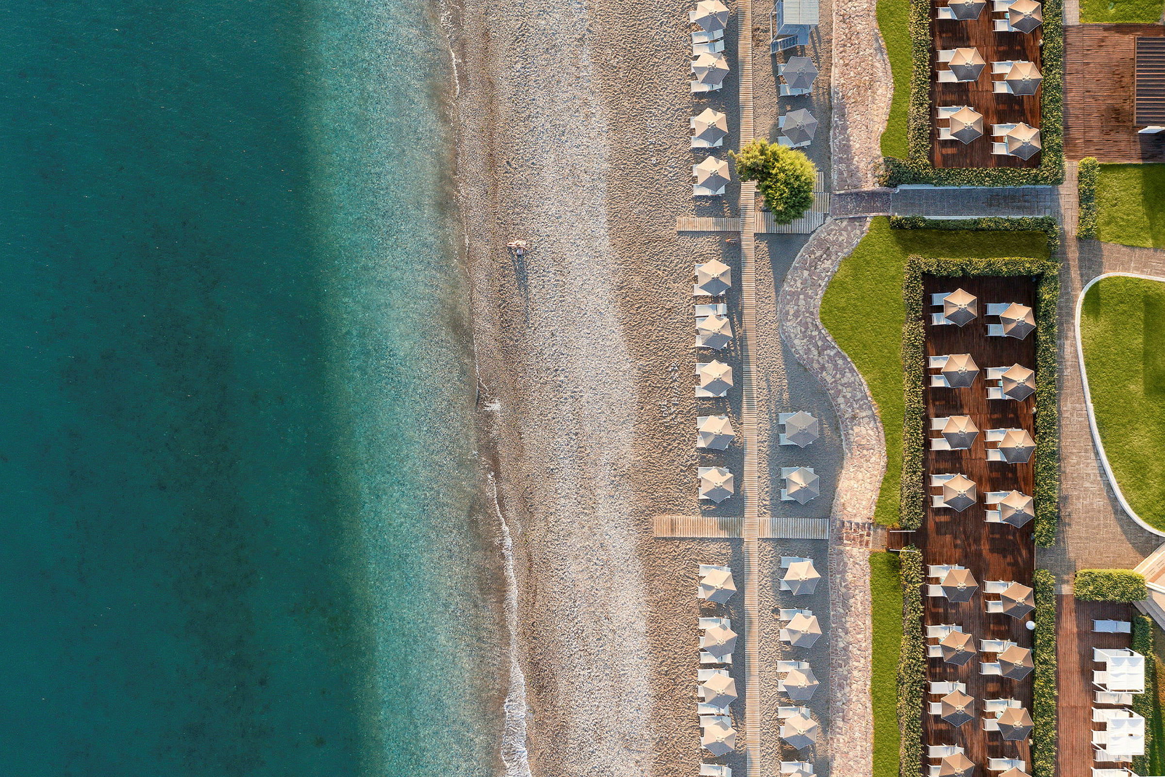 Flygfoto av en strand med klappersten och parasoller samt närliggande grönområden. (AI-genererad text)