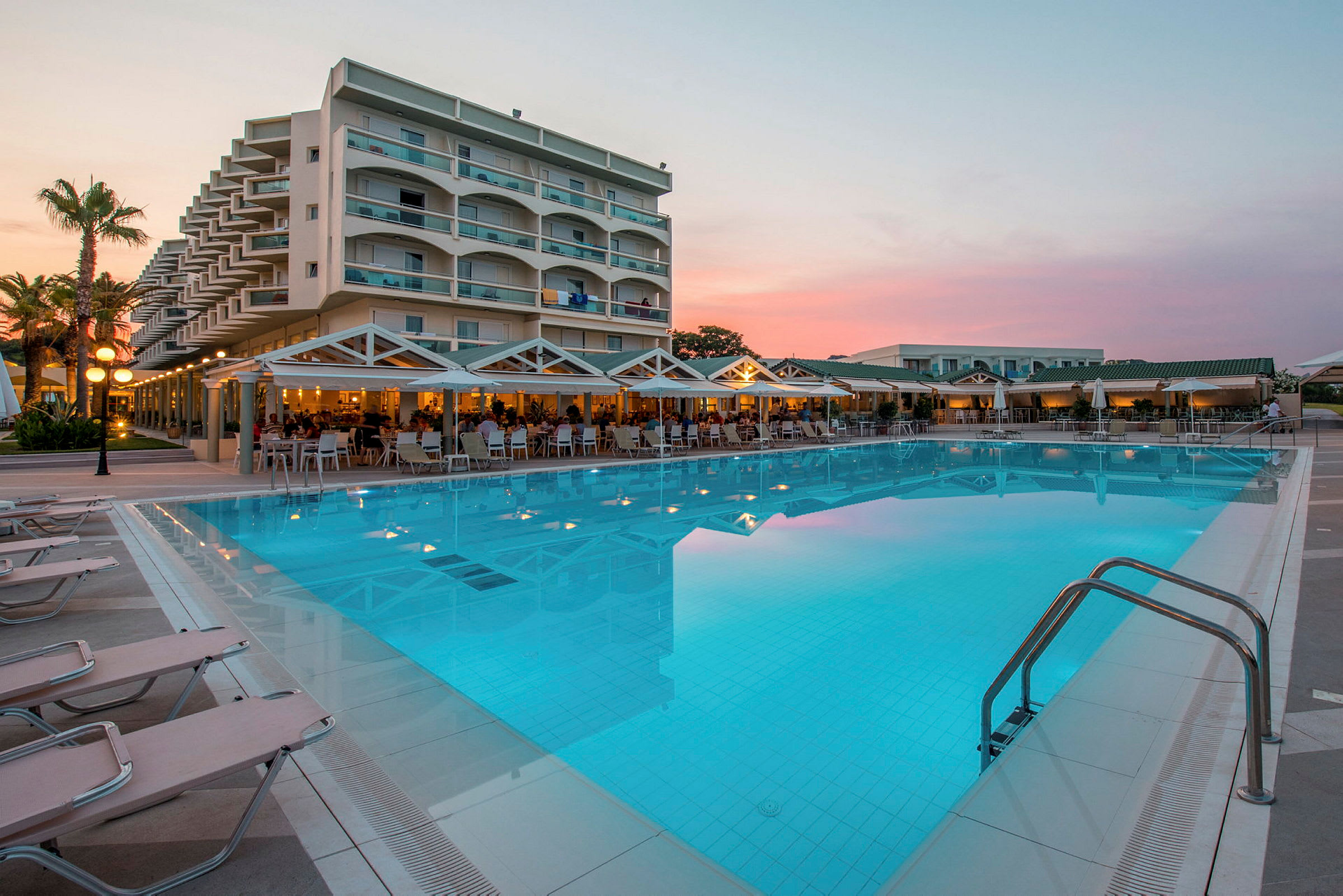 Hotellbyggnad med balkonger vid en stor swimmingpool omgiven av solstolar och en terrass med sittplatser. (AI-genererad text)