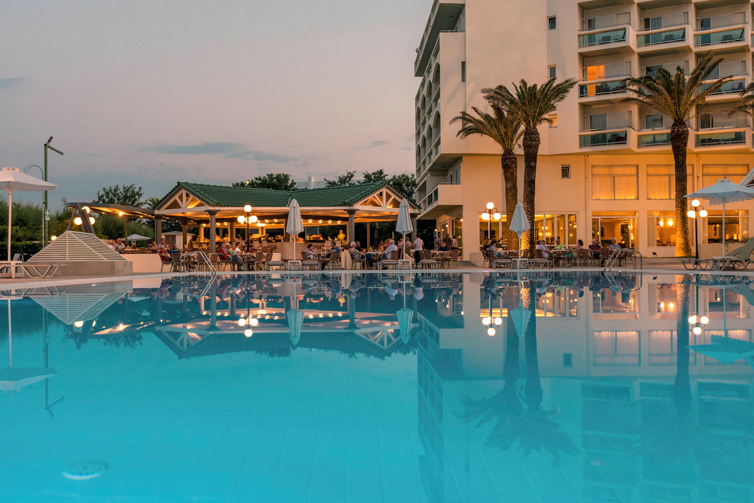 Hotellbyggnad med palmer och upplyst restaurang vid en stor pool. (AI-genererad text)
