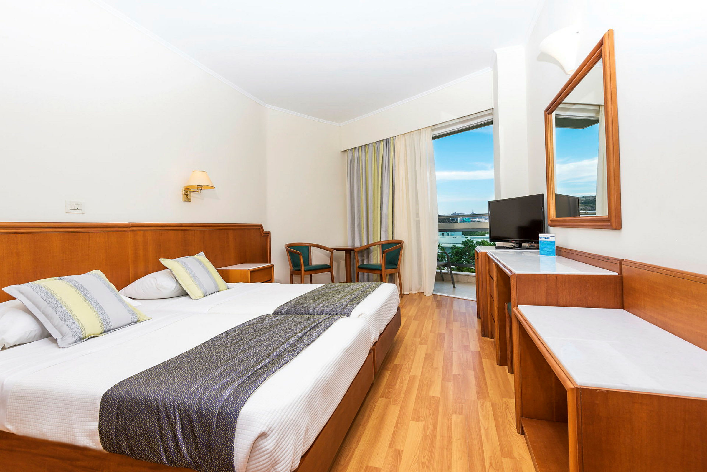 Hotellrum med dubbelsäng, skrivbord, TV och sittgrupp nära en balkong med utsikt. (AI-genererad text)