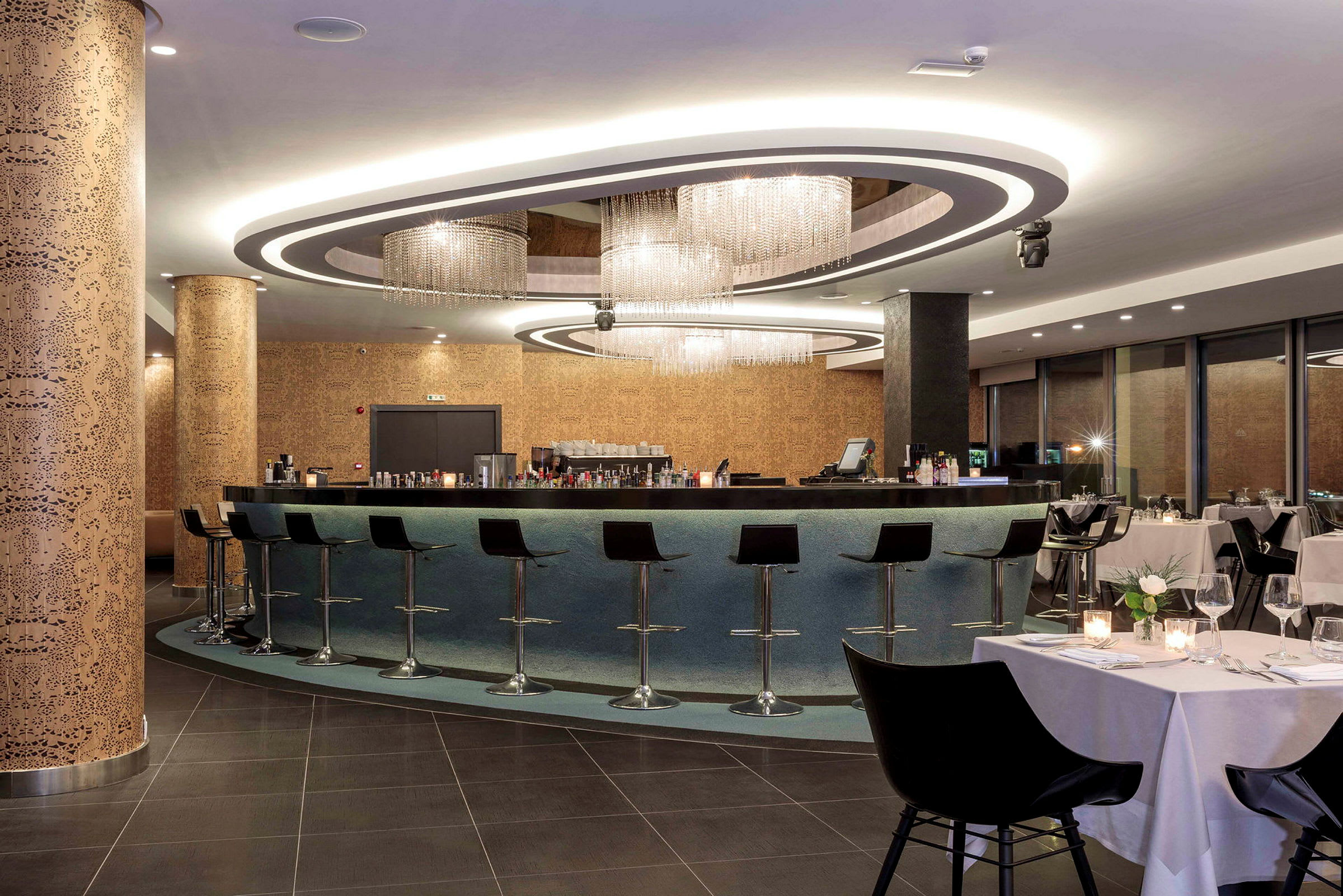 En modern bar med höga stolar och en elegant ljusdesign, omgiven av en restaurang med dukade bord. (AI-genererad text)