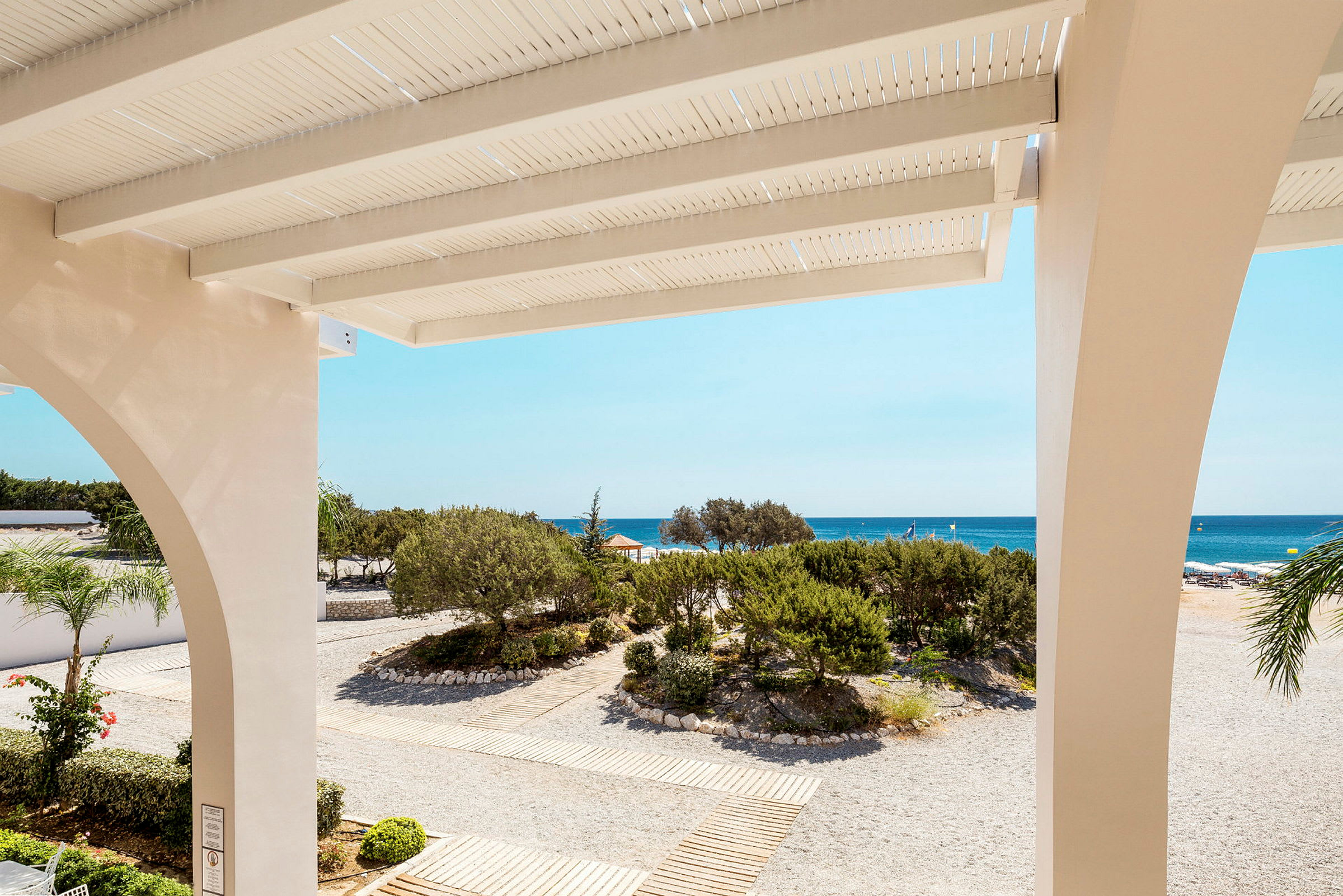 Utsikt från en terrass mot en strand med sand och grönska samt havet i bakgrunden. (AI-genererad text)