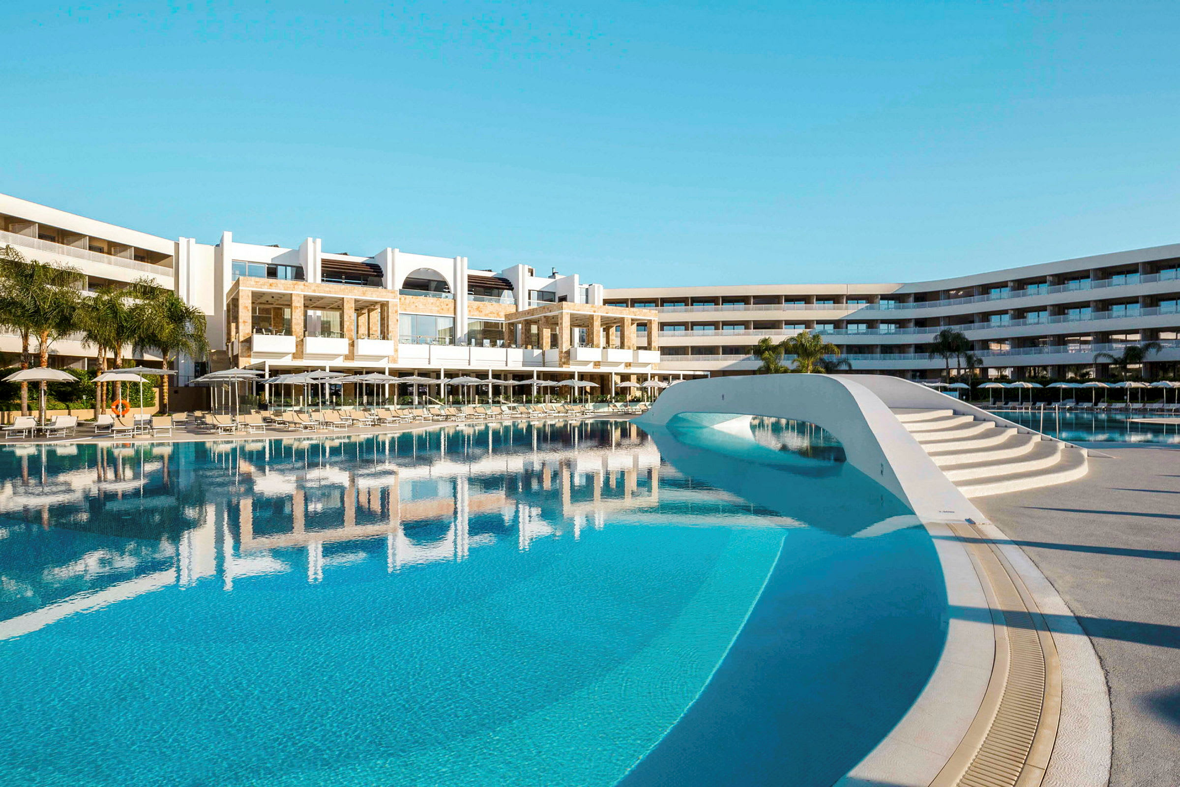Hotellbyggnad med ett stort poolområde och solstolar. (AI-genererad text)
