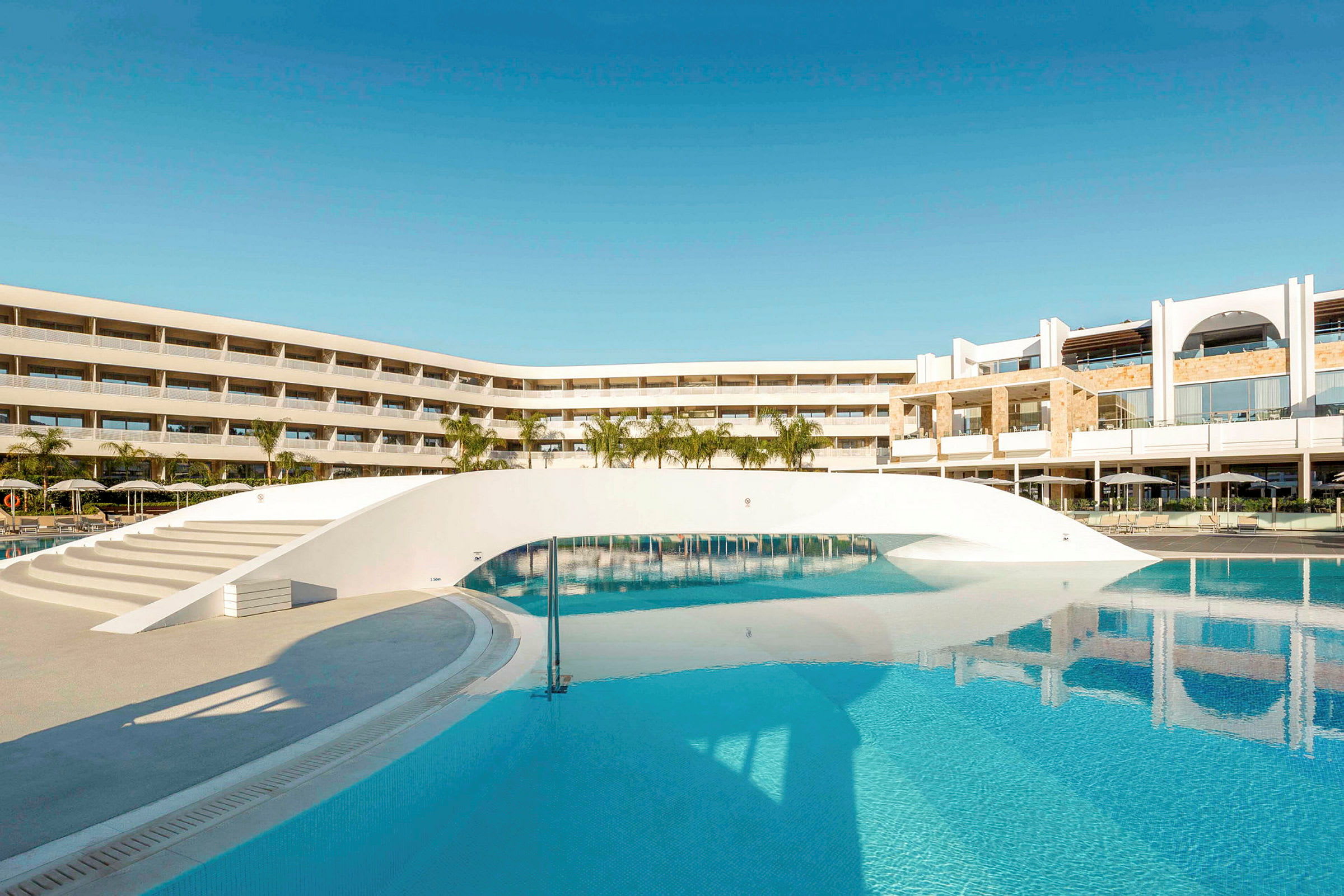 Ett modernt hotell med en stor pool och en vit bro över vattnet. (AI-genererad text)