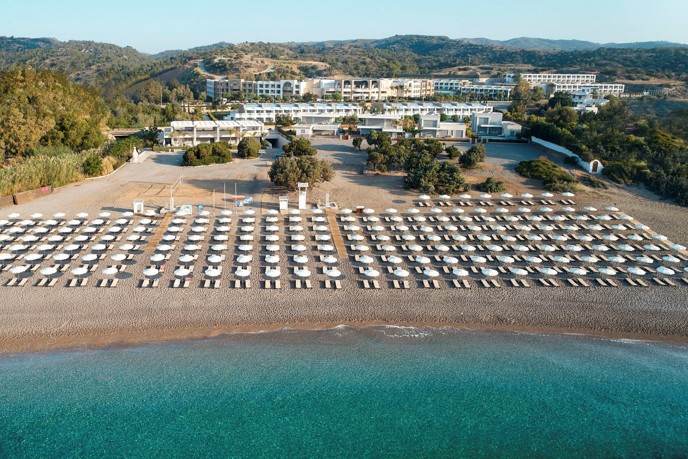 Solstolar och parasoller i flera rader på en sandstrand med turkost vatten, omgivna av grönska och byggnader i bakgrunden. (AI-genererad text)