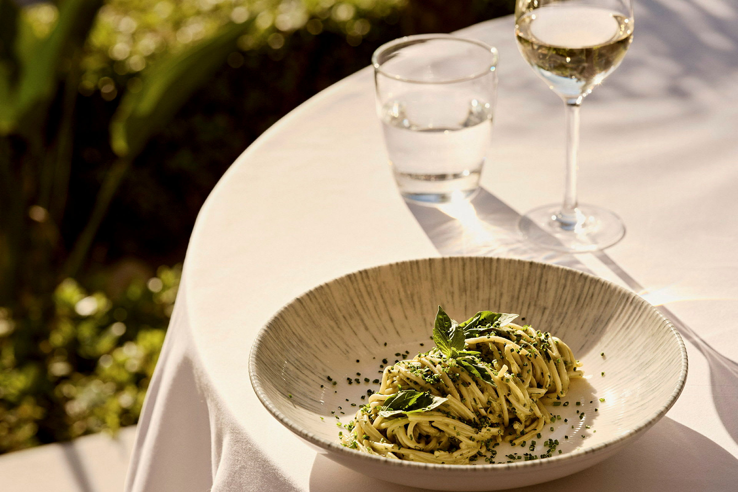 En tallrik med pasta dekorerad med örter, placerad på ett bord dukat med vit duk, bredvid ett glas vin och ett glas vatten. (AI-genererad text)