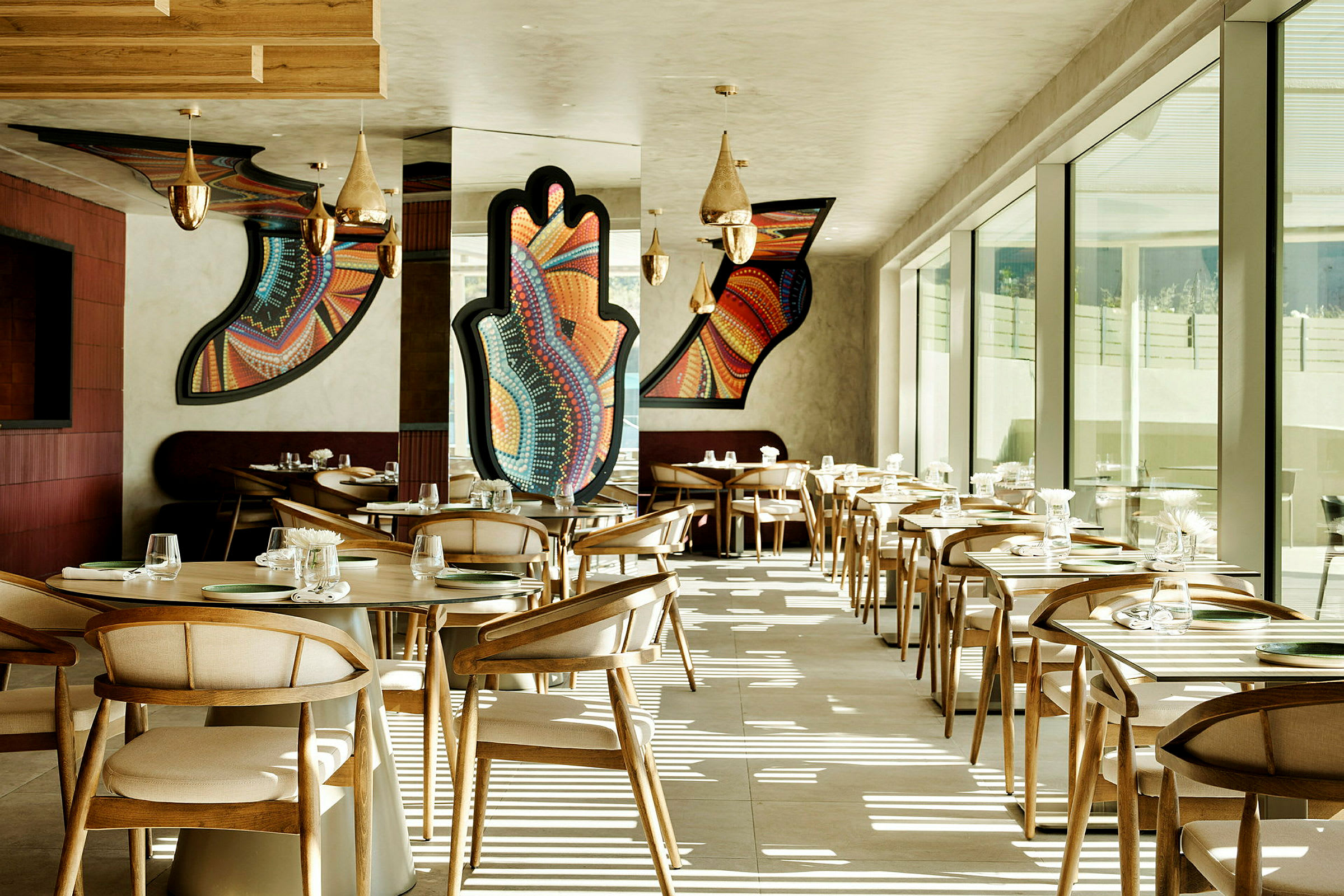 En modern restaurang med trästolar och bord. Väggarna har färgglada geometriska konstverk. Stora fönster släpper in naturligt ljus. (AI-genererad text)