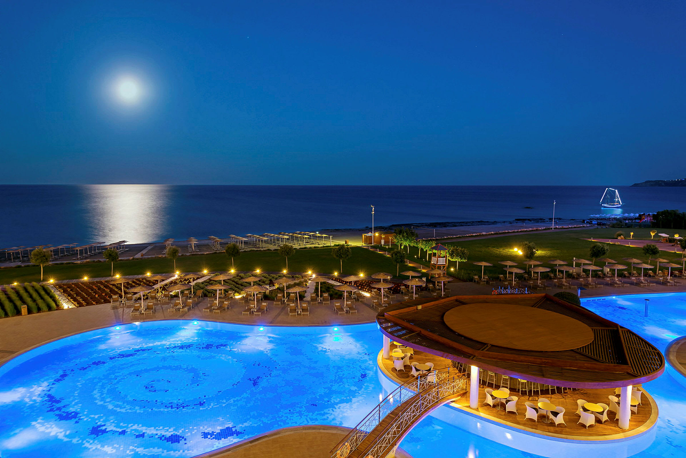 Kvällsvy över en upplyst pool vid en resort nära havet, med fullmåne och reflektioner på vattnet. (AI-genererad text)