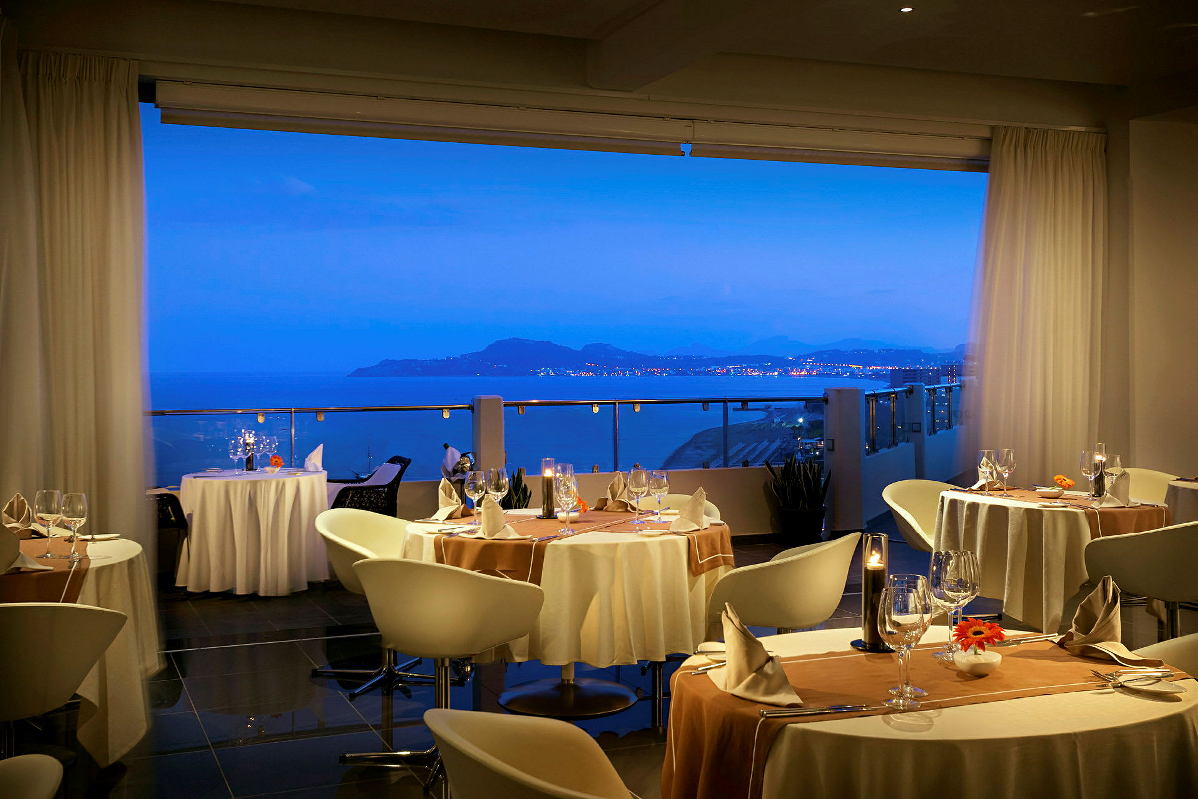 Utsikt mot en havsvik med avlägsna ljus på land från en elegant restaurang interiör. (AI-genererad text)