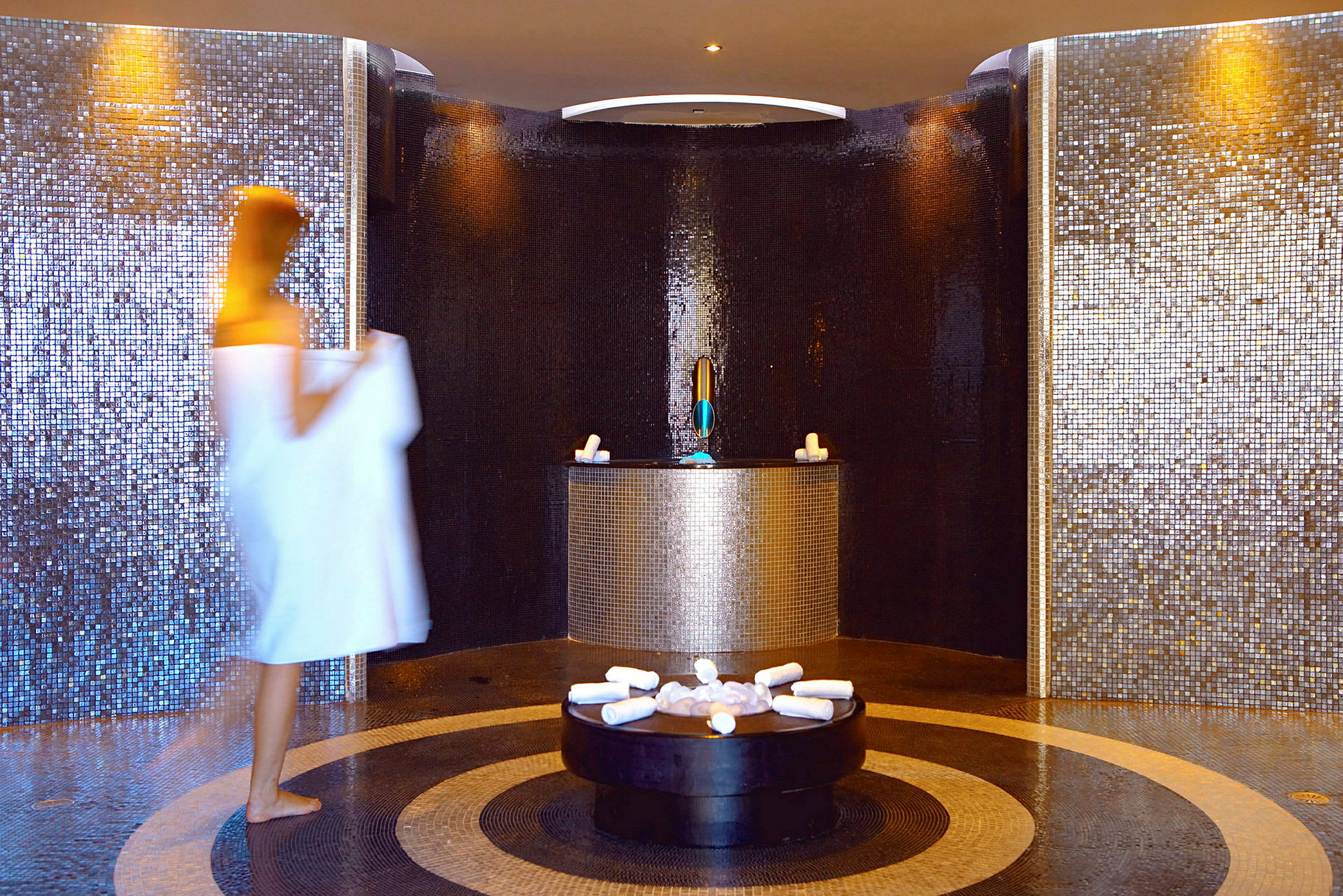 En person i vit handduk går genom ett spa med mosaikväggar och cirkulärt golv. (AI-genererad text)