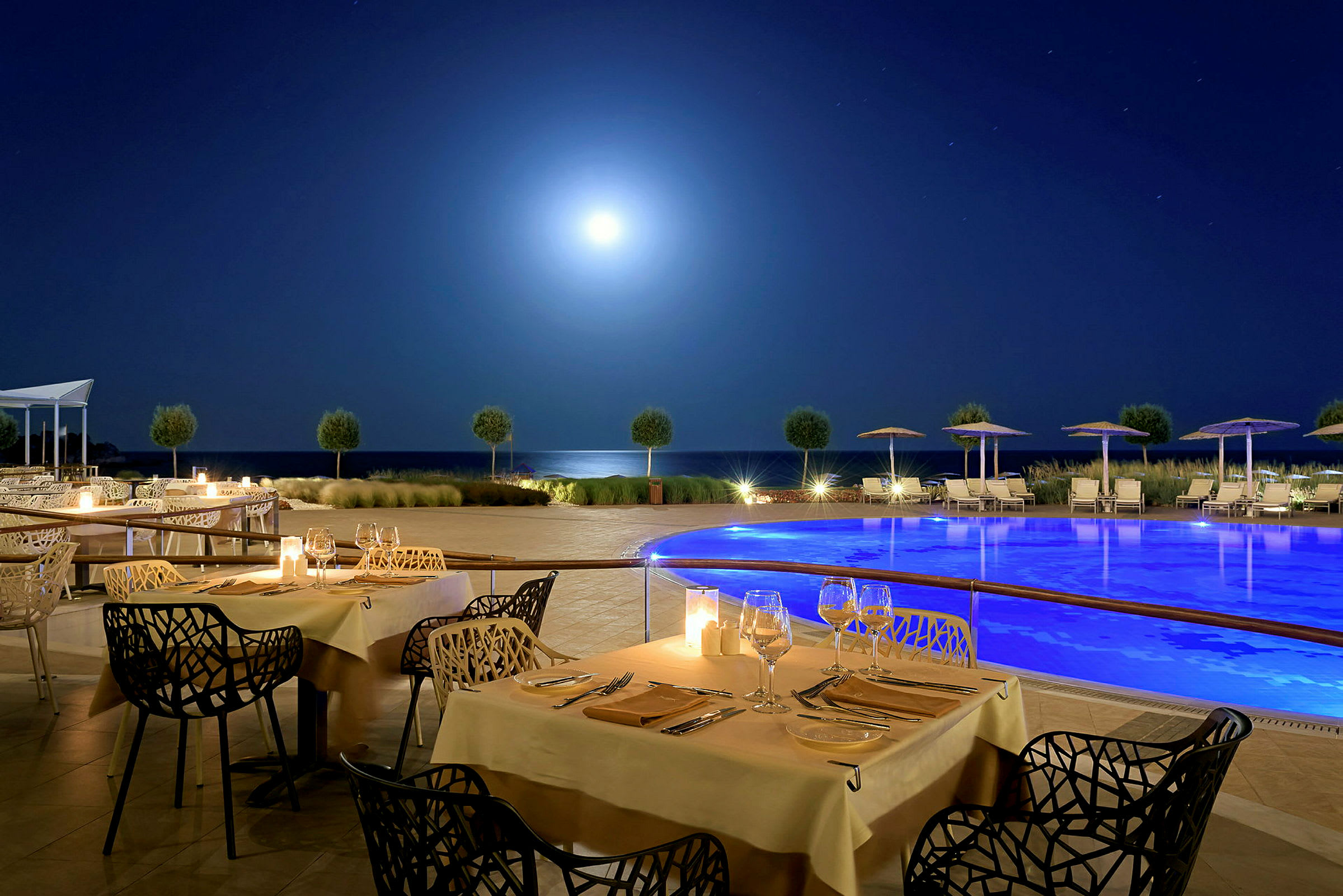 En utomhusrestaurang med dukade bord och en pool i närheten under en stjärnklar natthimmel. (AI-genererad text)