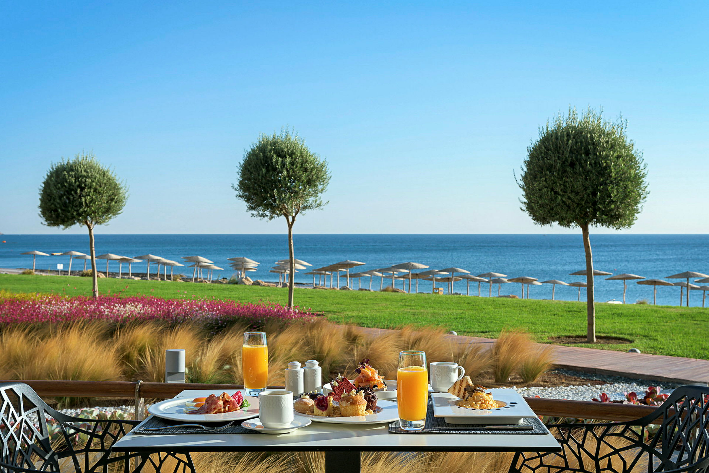 Frukost serverad vid ett bord med utsikt över havet, grönska och parasoller. (AI-genererad text)
