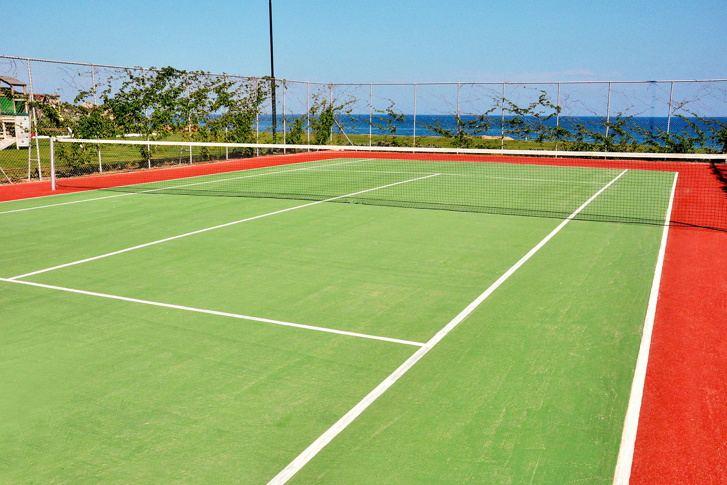 En tennisplan med grön yta och röda kanter, omgiven av ett nätstängsel och grönska, med havet synligt i bakgrunden. (AI-genererad text)