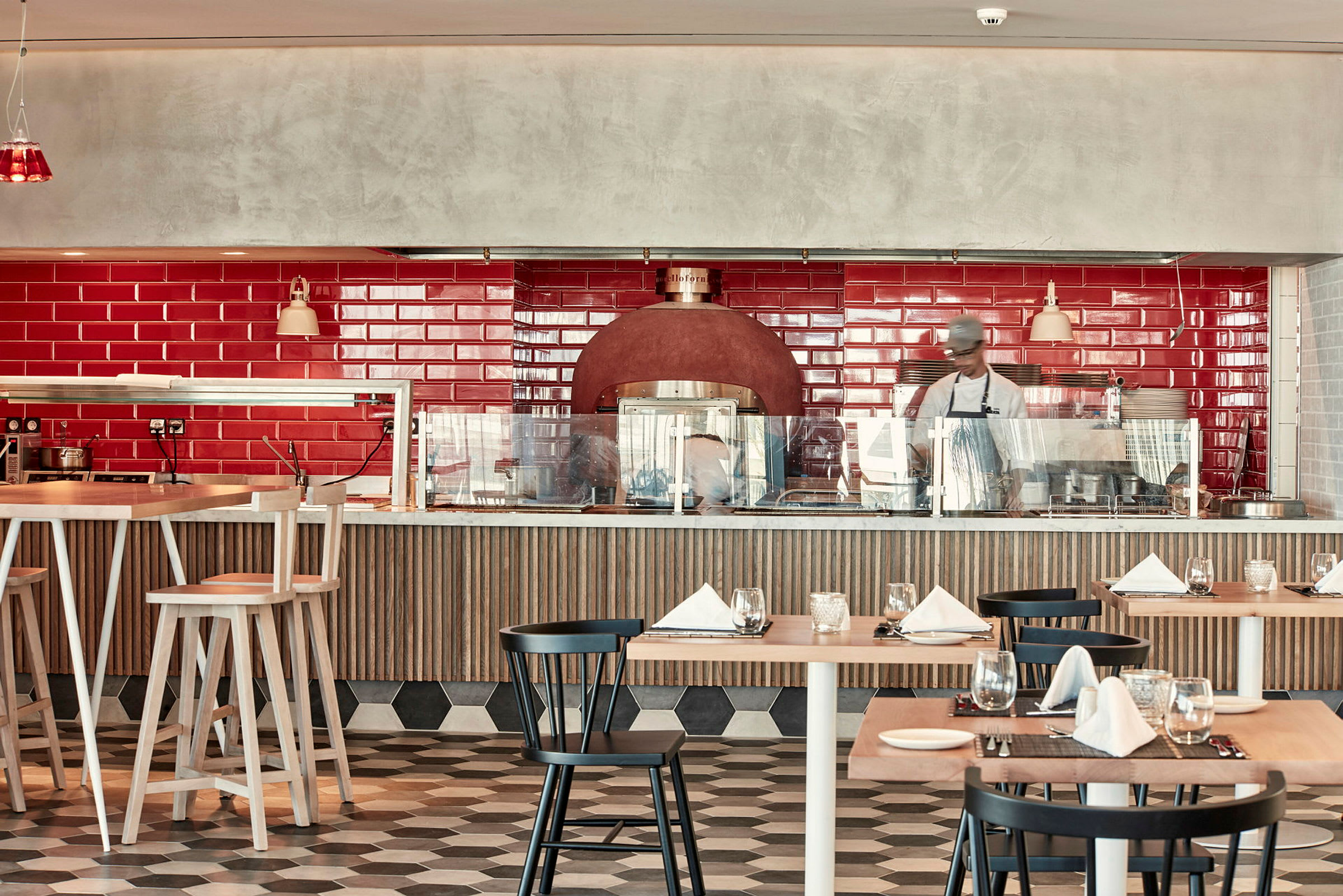 En restauranginteriör med röd kaklad vägg, matbord och stolar samt en vedugn. (AI-genererad text)