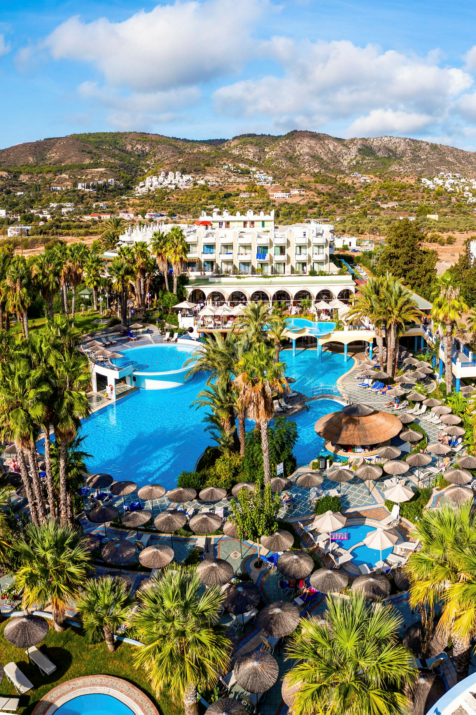 Bild av ett hotellområde med pooler, parasoller och palmer, beläget vid foten av berg. (AI-genererad text)
