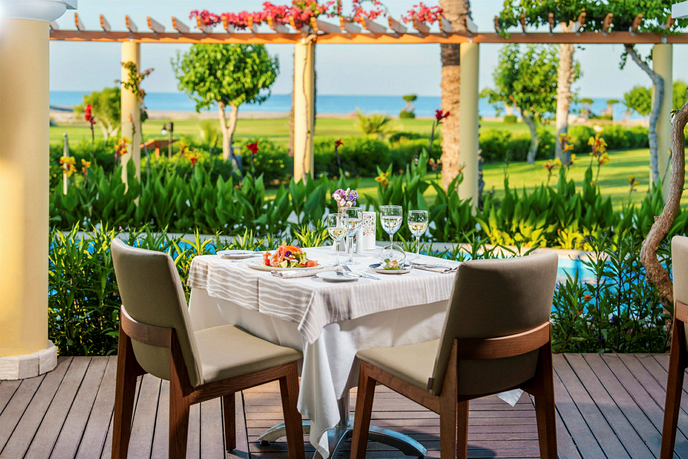Två matstolar vid ett dukat bord stående på en terrass med utsikt över en trädgård och havet. (AI-genererad text)