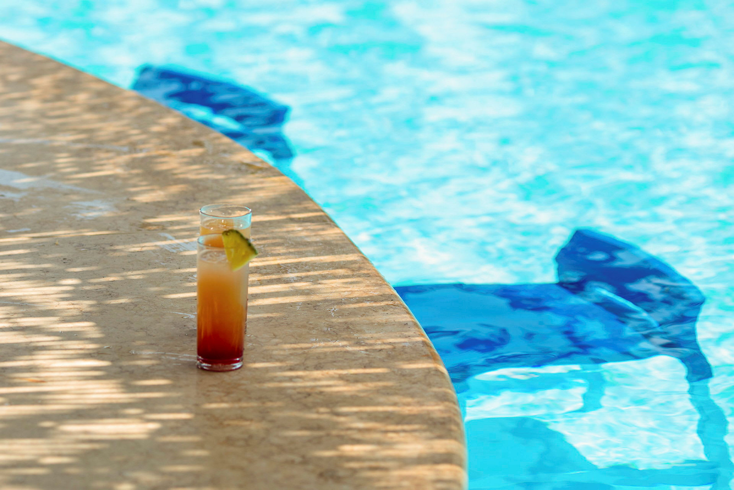 Ett glas med en färgglad drink står på en stenkant bredvid en swimmingpool. (AI-genererad text)