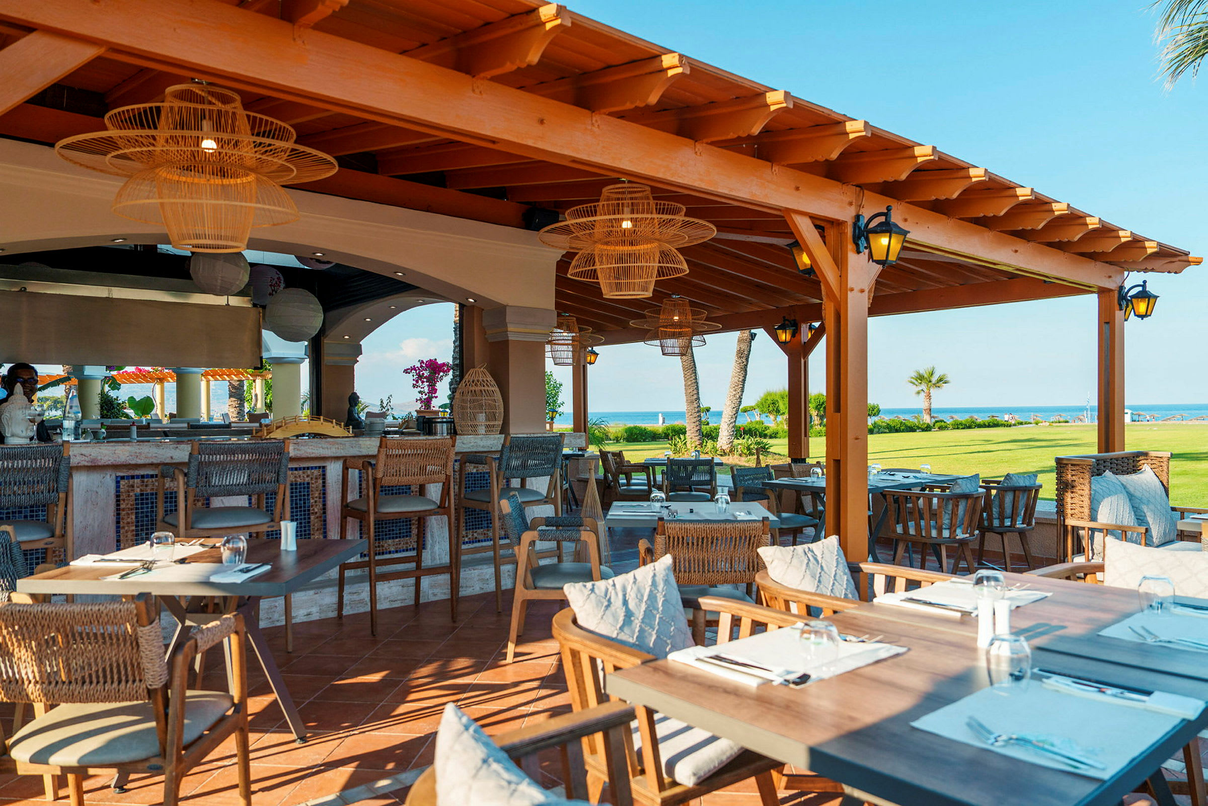 En restaurang med uteservering vid havet, inredd med trädetaljer och rottingmöbler. (AI-genererad text)