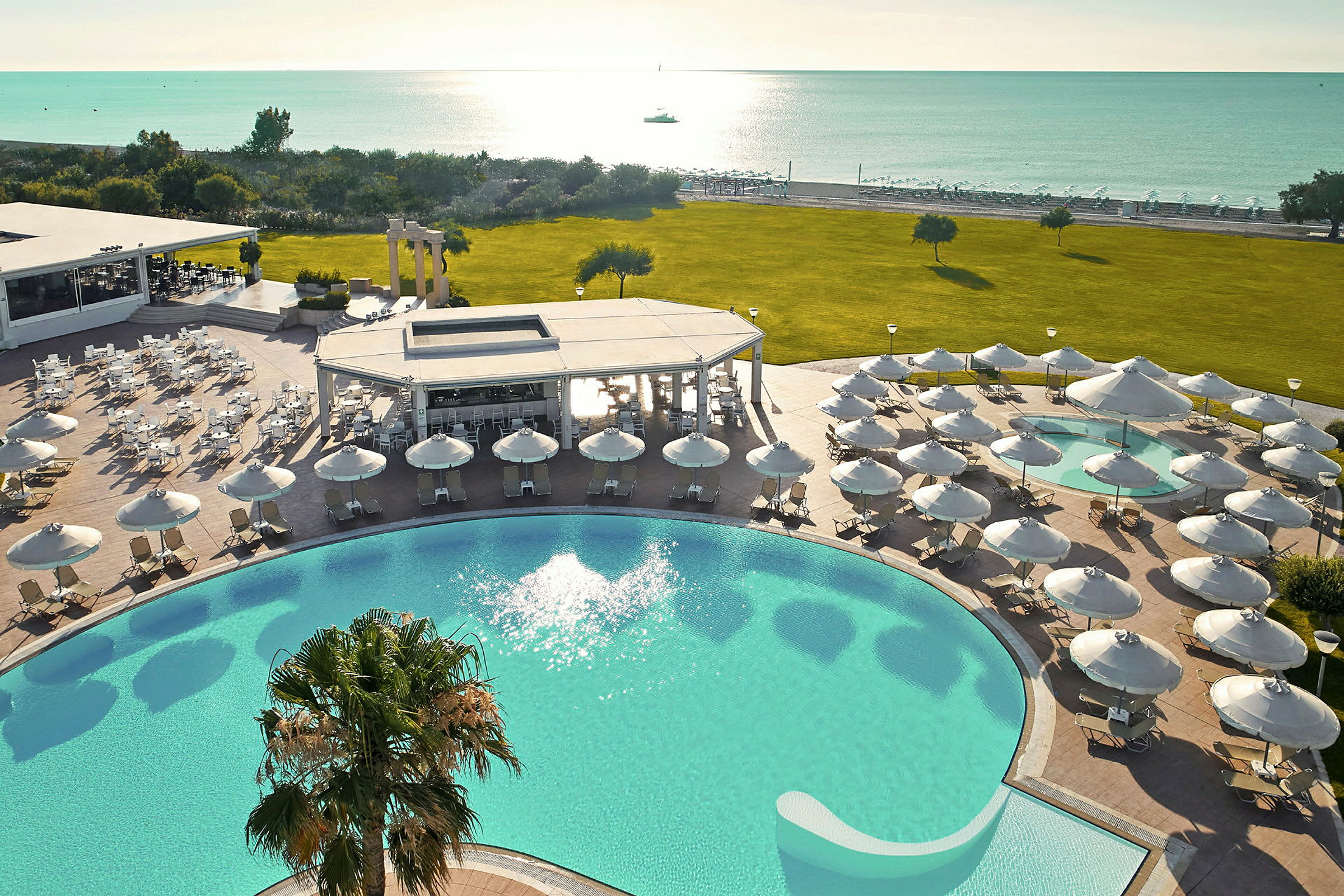 En vy över en solig resort med pool, parasoller, grön gräsmatta och utsikt över havet. (AI-genererad text)