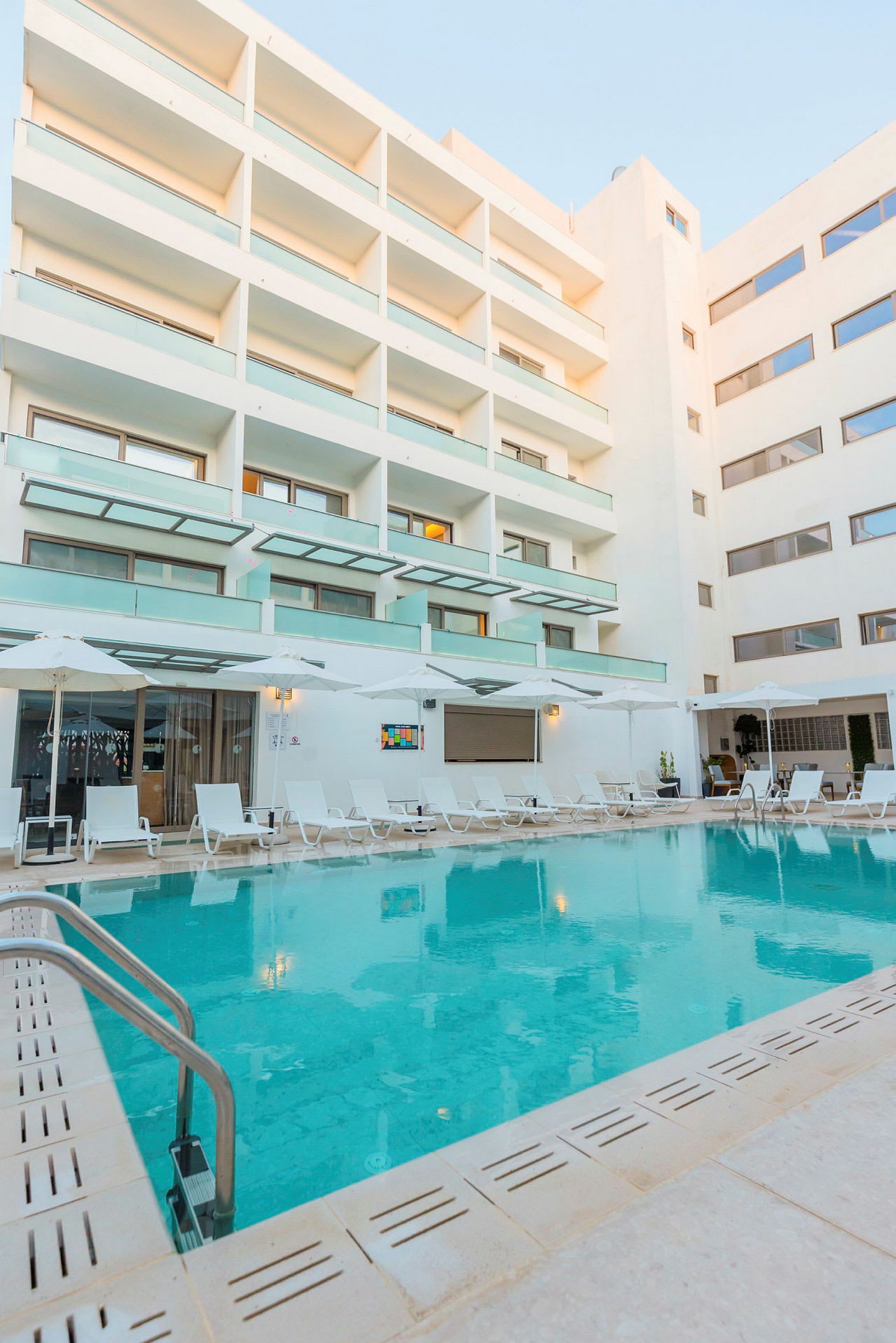 En hotellpool omgiven av solstolar och ett modernt hotellkomplex. (AI-genererad text)