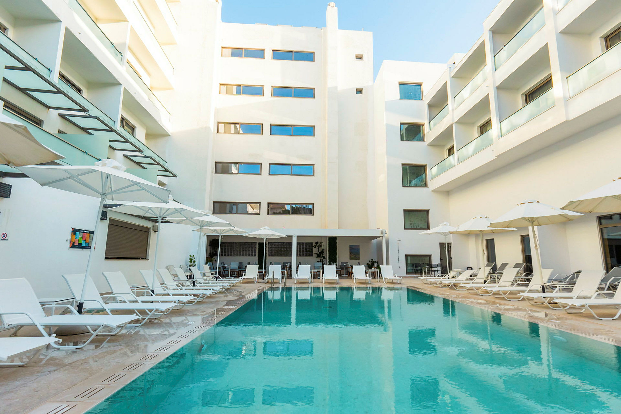 Bild av ett hotellområde med en pool omgiven av solstolar och parasoller ovanför. Bakgrunden visar vita byggnader med balkonger. (AI-genererad text)