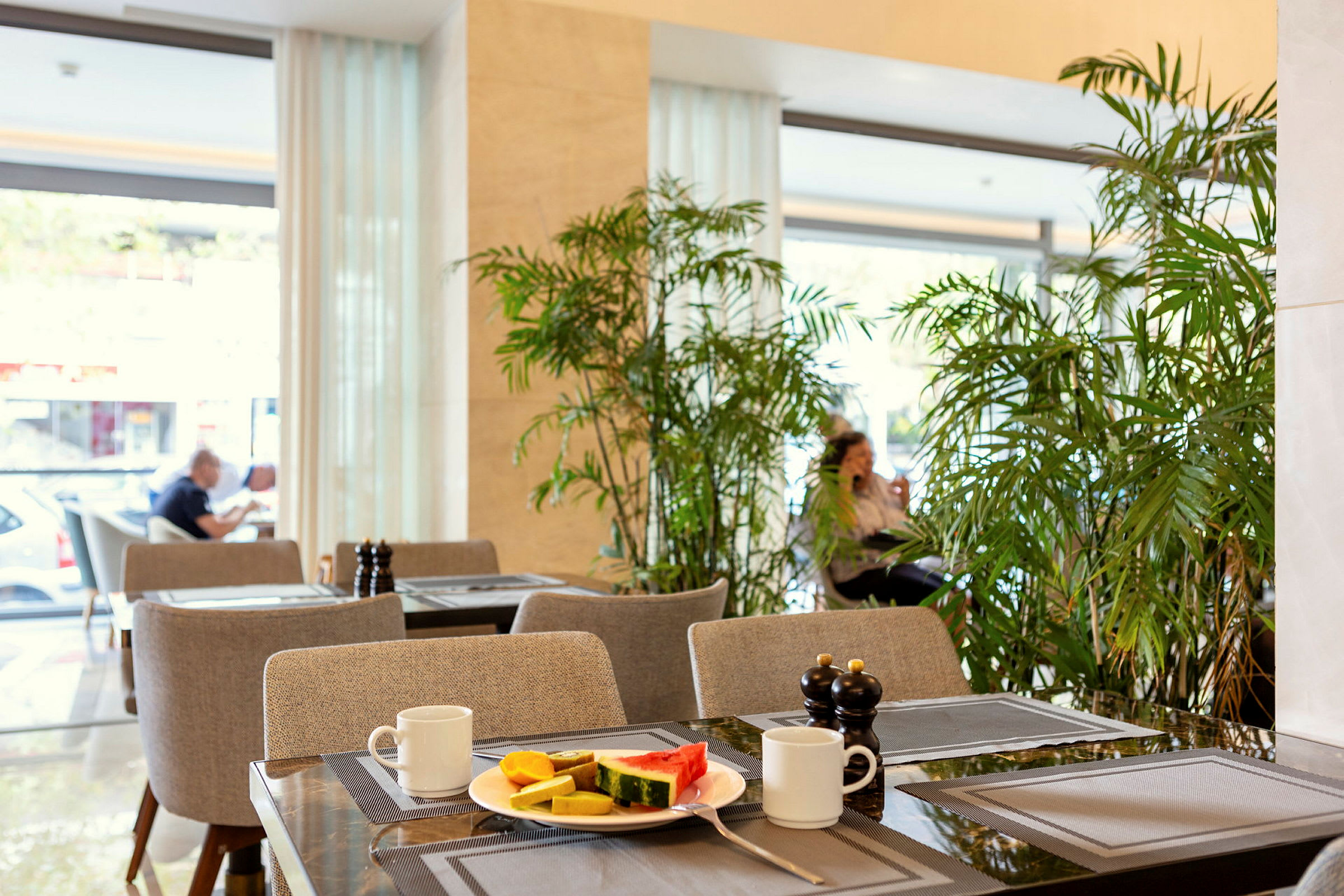 Ett café med bord som är dukade med tallrik innehållande frukt, koppar och kryddbehållare, omgivet av grön växtlighet. (AI-genererad text)