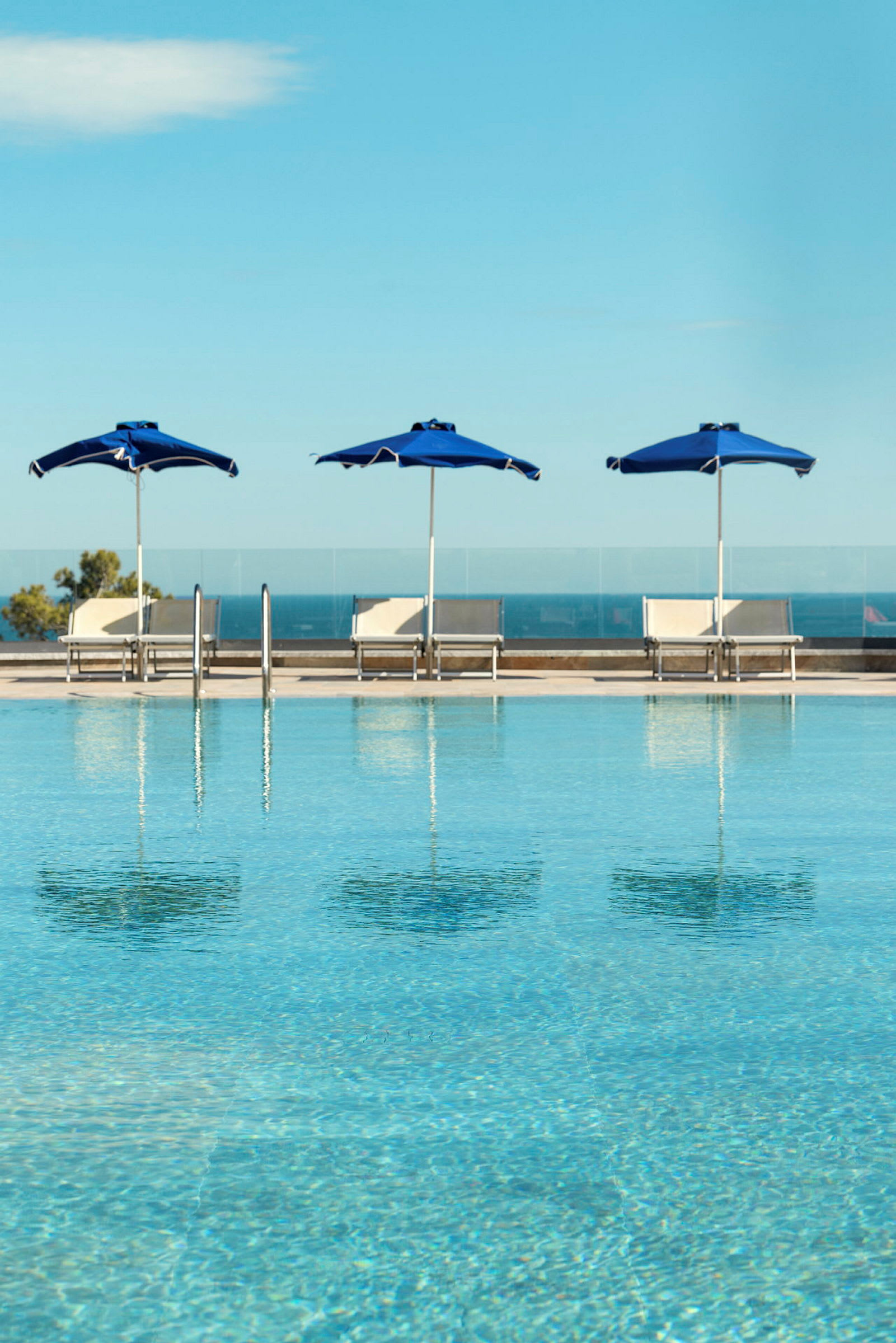 En pool omgiven av blå parasoller och solstolar med utsikt mot havet. (AI-genererad text)