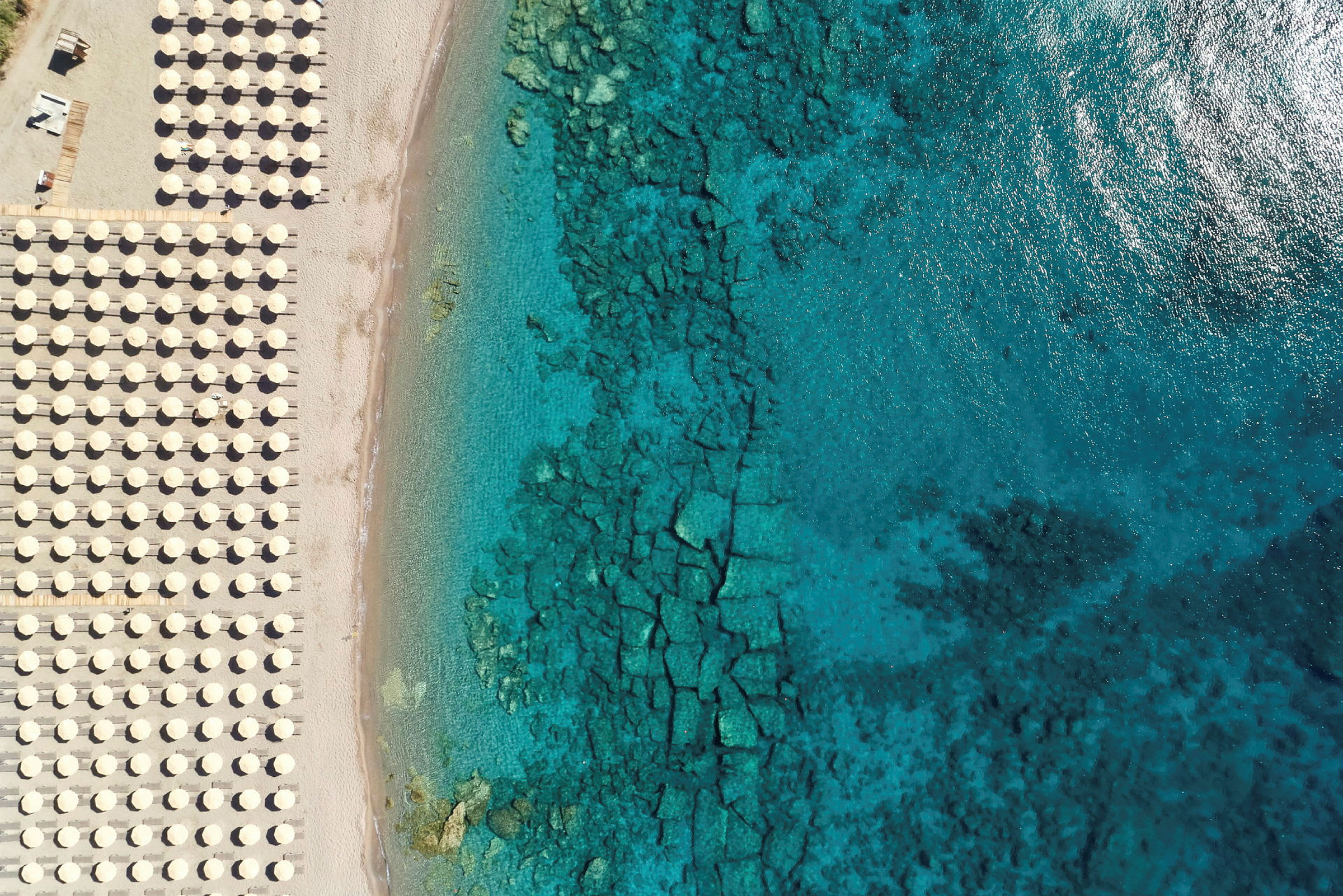 Flygfoto över en strand med solstolar och parasoller på vänster sida och klart blått hav på höger sida. (AI-genererad text)